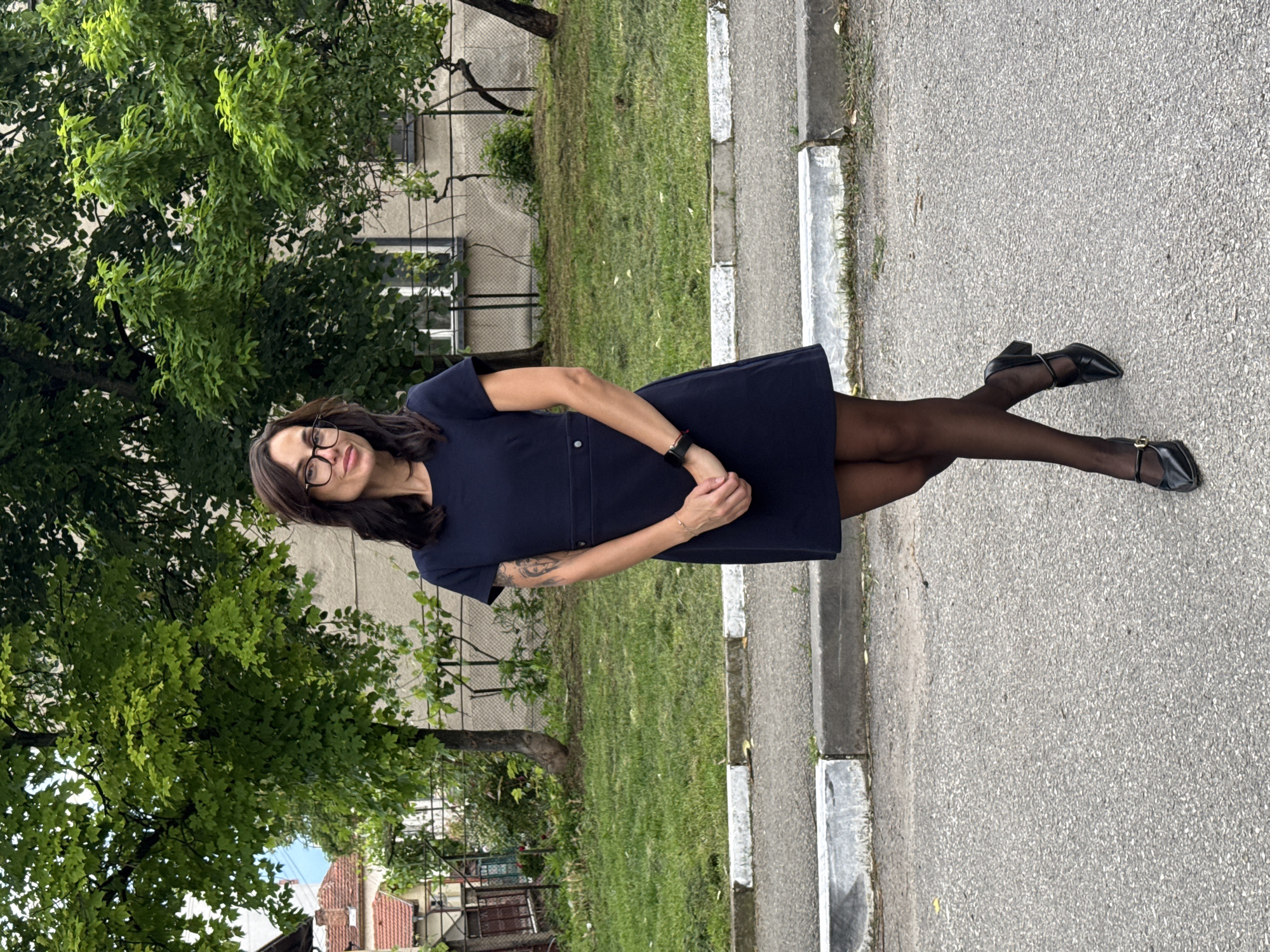 A medium shot captures a woman in a dark blue dress, tights, and black shoes standing on a sidewalk. She wears glasses and has a watch on her left wrist. Her arms are crossed in front of her. To her left are lush green trees and behind her is a building with barred windows and a grassy yard. To her right is the gray sidewalk and a street. The background shows part of Pleven, Bulgaria.