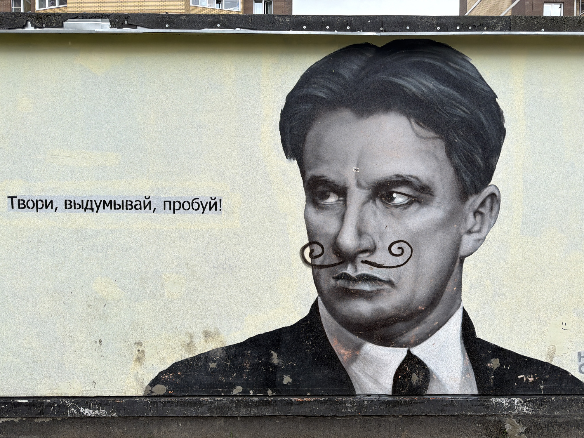 A mural in an unknown city features a portrait of a man with a dark mustache. The man is facing right and wearing a suit and tie. To the left of the portrait is the text "Твори, выдумывай, пробуй!". The mural is on a yellow wall with a dark trim at the bottom and a building in the background.