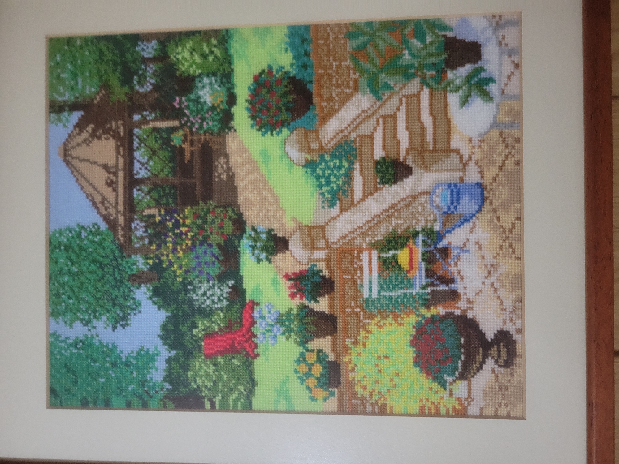 The image depicts a framed cross-stitch embroidery of a garden scene. The scene features a gazebo in the background, a pathway leading to it, and various potted plants and flowers. In the foreground, there are steps leading down to a patio area with a table and chairs, a watering can, and a large potted plant. The overall impression is of a tranquil, well-maintained garden. The location of Belaazyorsk, Belarus, is not discernible from the image itself, as there are no identifying landmarks or text present. The embroidery is enclosed in a white mat and a wooden frame.