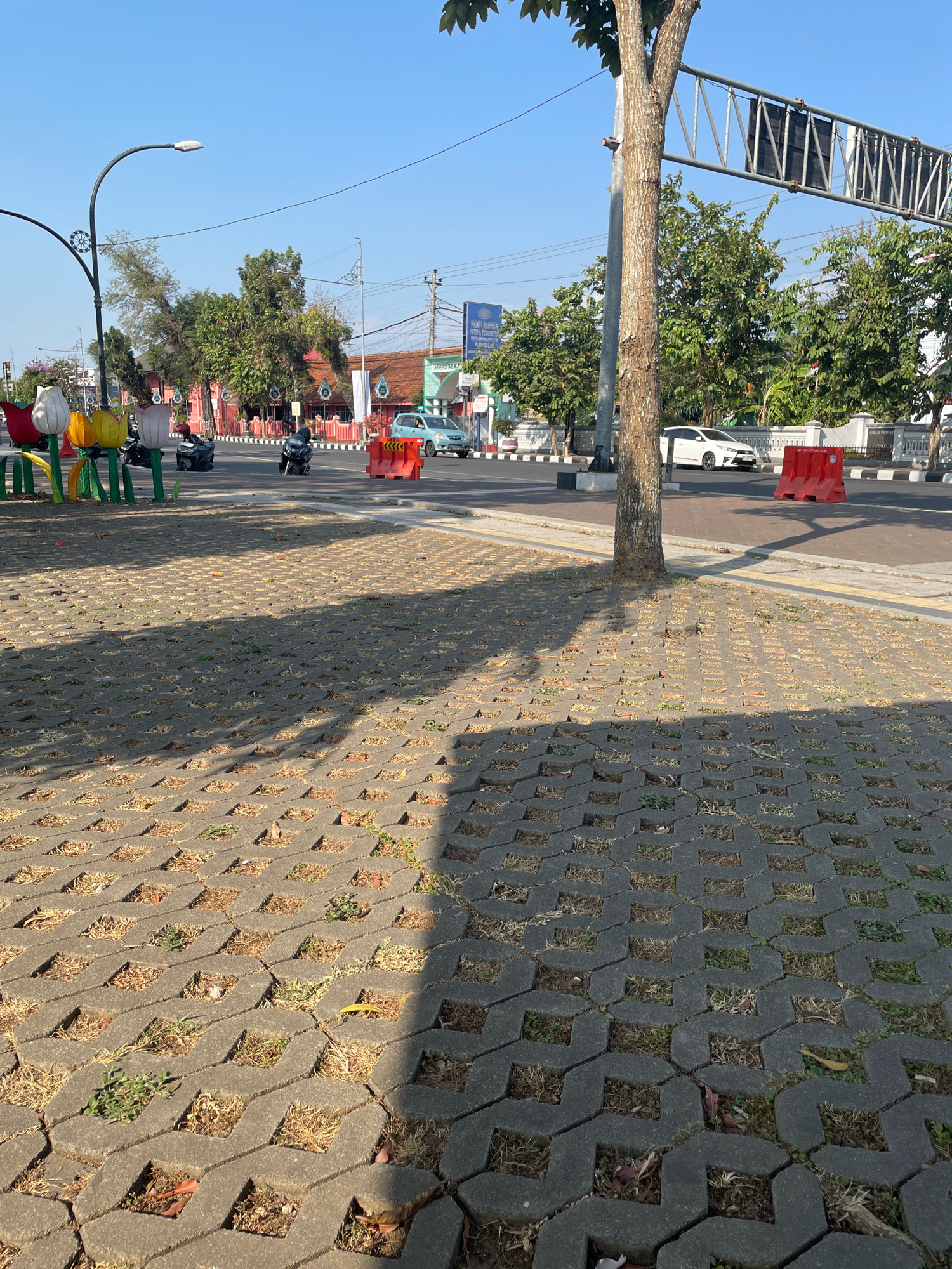 The image depicts a street scene in Purworejo, Indonesia, under a clear blue sky. In the foreground, a permeable pavement made of interlocking concrete blocks is visible, with patches of dry grass and some fallen leaves within the gaps. A tree trunk is prominently positioned in the mid-ground, casting a shadow across the pavement.
The street itself features a white car and several motorcycles in motion. On the roadside, there are decorative tulip-shaped objects, some colorful plastic chairs, and concrete barriers painted orange. Buildings with red tile roofs line the opposite side of the street, and various trees with green foliage are scattered throughout the scene. Overhead, a traffic signal structure with several black panels is visible. A lamppost with a modern design stands near the left side of the frame. The environment appears to be an urban or suburban setting with ongoing traffic and pedestrian-related infrastructure.