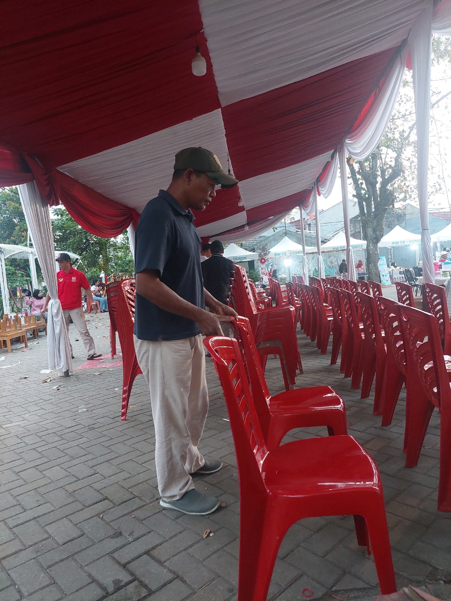 An outdoor event is being held under a red and white striped tent. The ground is paved with rectangular bricks. In the foreground, a man wearing a dark blue polo shirt and light-colored pants is standing and appears to be arranging red plastic chairs. Behind him, a row of red plastic chairs is neatly arranged. Further in the background, another man in a red shirt and light pants is walking. Other people are visible in the distance, as well as white tents and trees. The scene suggests preparations for an event, possibly a community gathering or a local celebration in Depok, Indonesia.
