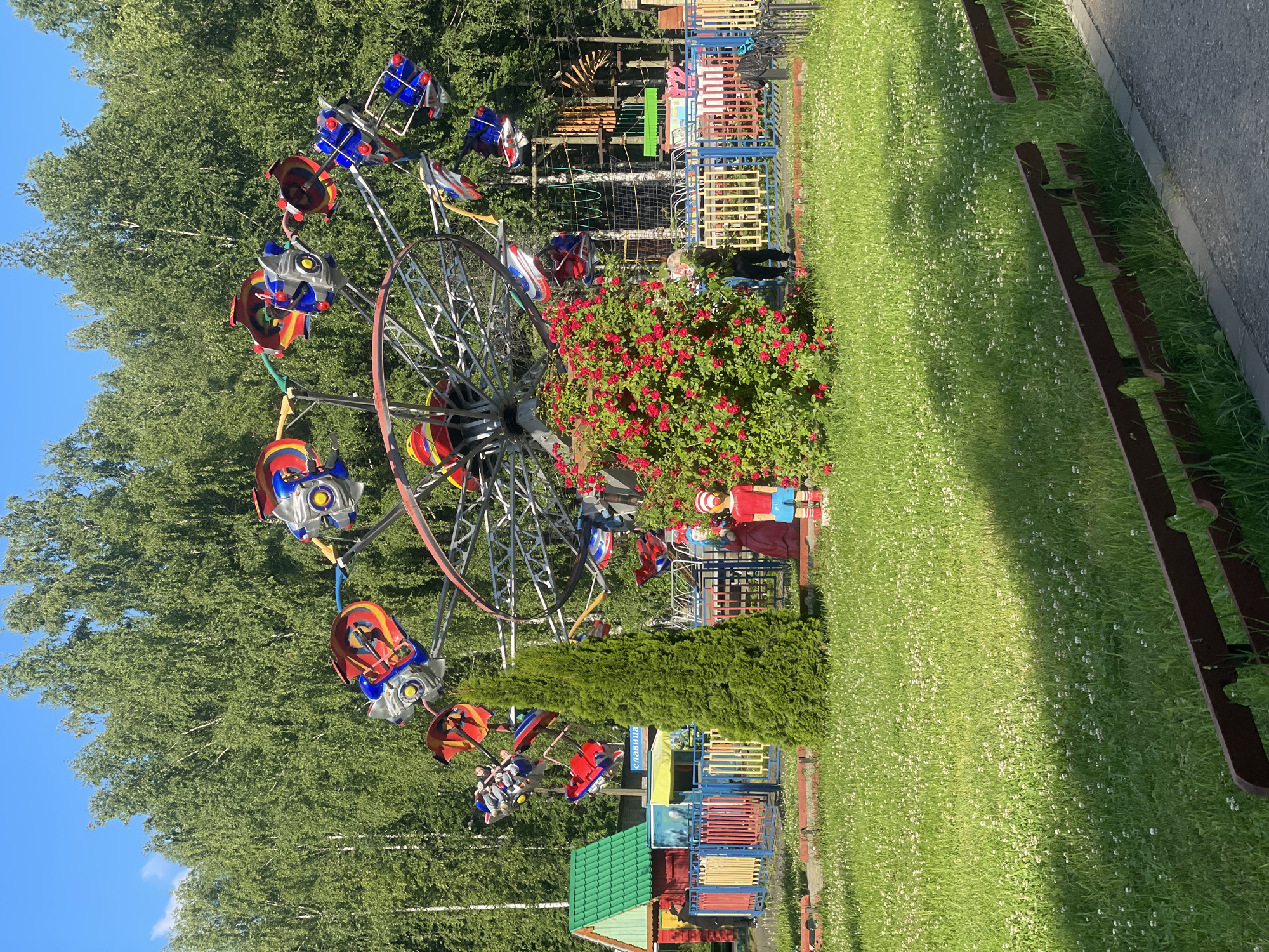 An outdoor amusement park in Chaikovsky, Russia, features a colorful spinning ride with individual pods. A young boy in a red shirt and blue shorts stands near a large bush with red flowers. In the background, a small red and green building with a colorful fence is visible, along with a tall evergreen tree. The park is set against a backdrop of lush green trees under a bright blue sky. The ground is covered in green grass dotted with small white wildflowers. A wooden fence runs along the bottom of the image.