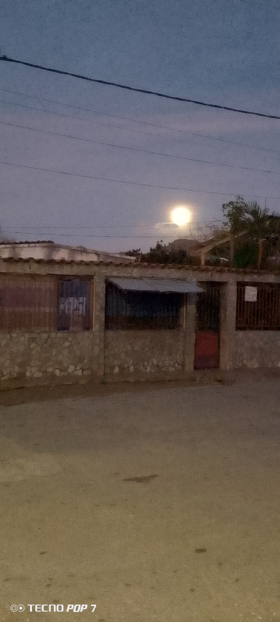 The image shows a building in Altagracia, Venezuela, at dusk or night. The building is single-story with a stone facade and a corrugated metal roof. There are several window openings, some covered with metal screens. One section has a red door. A Pepsi advertisement is visible on one of the window sections. Power lines run across the top of the frame. In the sky, a bright moon or light source is visible behind the building. The street in front of the building appears unpaved.