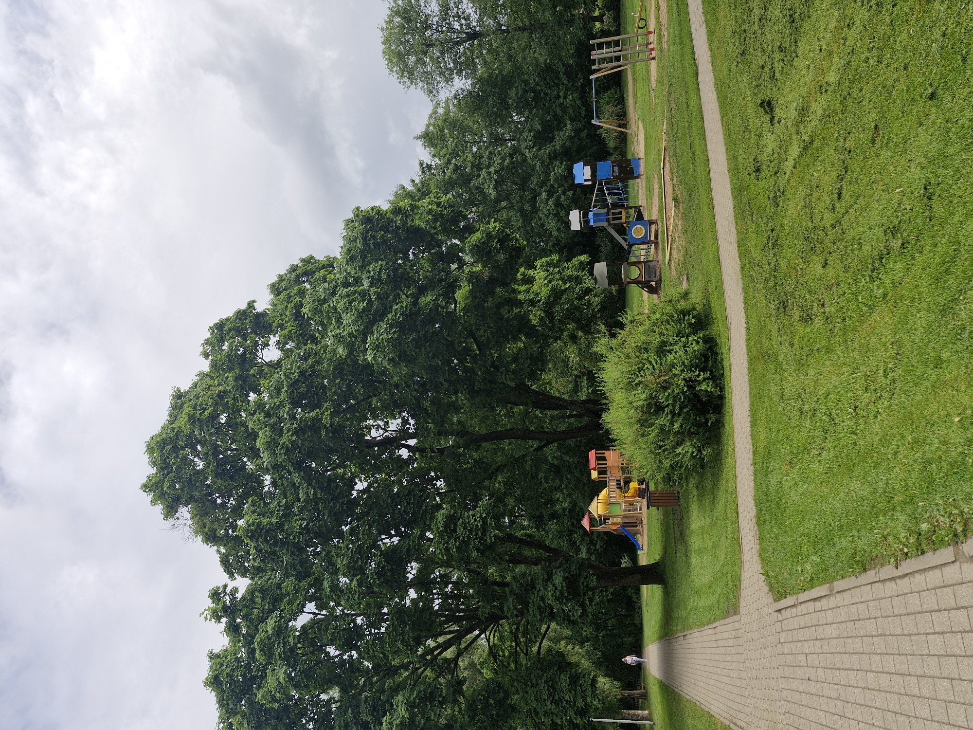 An outdoor daytime scene depicts a paved walkway on the left, leading into a park with lush green grass and mature trees. A single pedestrian, a person wearing a light-colored top and dark pants, walks away from the camera on the walkway. To the right of the walkway, a brightly colored children's playground is visible, featuring slides, climbing structures, and swings. In the background, dense green foliage forms a natural backdrop. The sky is overcast with gray clouds.
