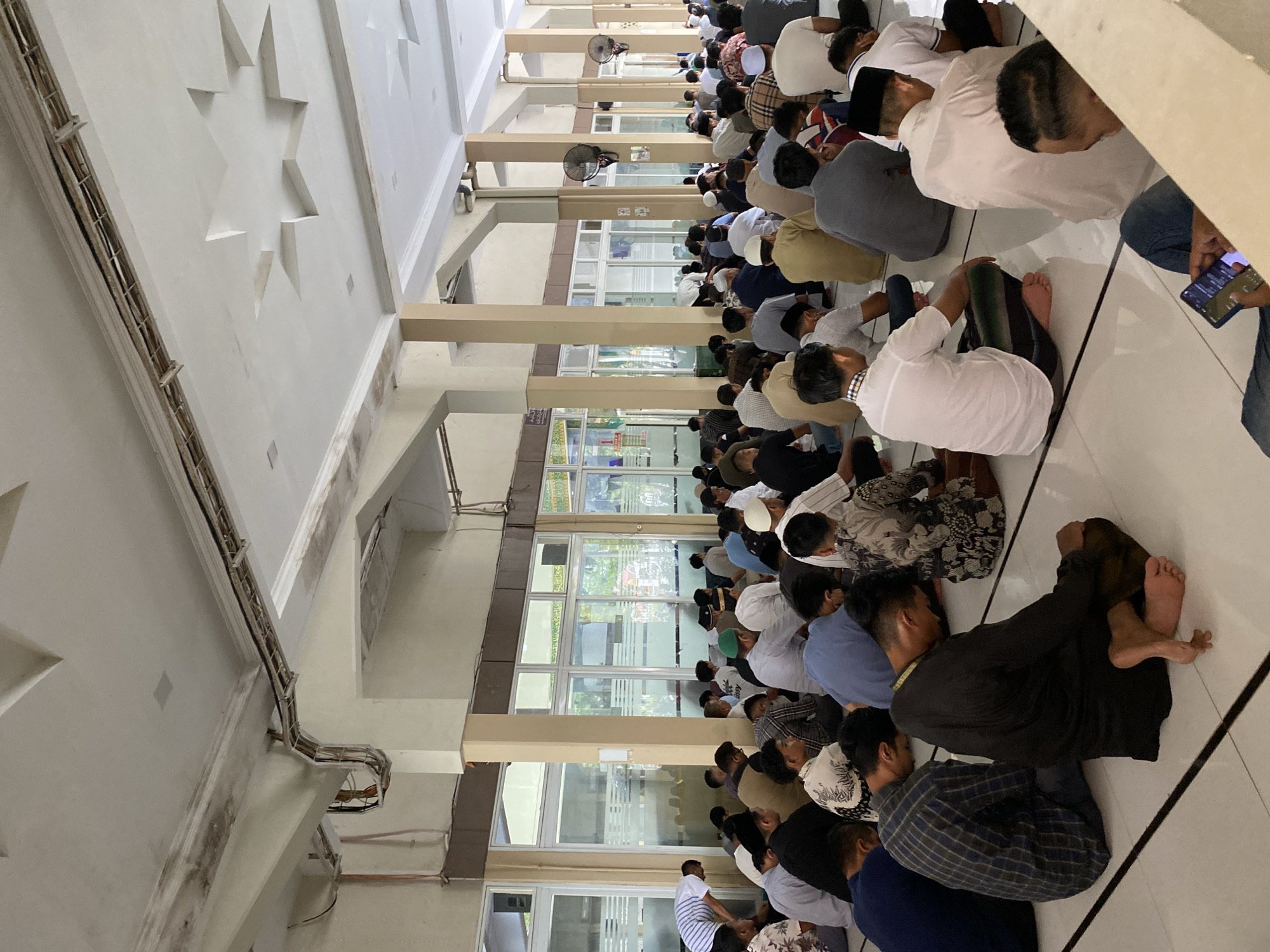 The image depicts a large group of men sitting on a tiled floor inside a mosque in Lhokseumawe, Indonesia. The men are facing the same direction, suggesting they are engaged in prayer or a religious gathering. They are dressed in various clothing, including long shirts, trousers, and head coverings, typical attire for Muslim men. Some men are leaning forward, while others are sitting upright. The environment appears to be a spacious hall with columns, large windows with reflective glass, and a white ceiling with decorative star-shaped elements. There are also electrical conduits and fans visible on the ceiling and walls. The lighting is diffuse, suggesting it is daytime, with light coming in through the windows. The overall atmosphere is one of solemnity and community.