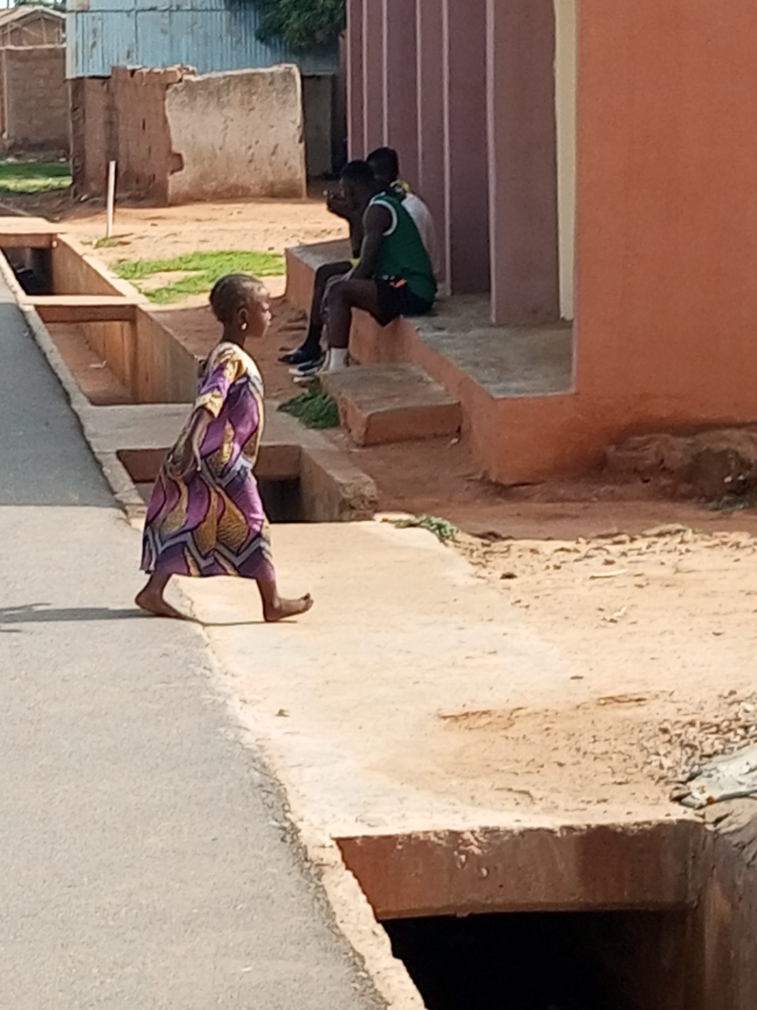 In Tempure, Nigeria, a young child walks barefoot across a paved area. The child is wearing a vibrantly patterned purple, yellow, and blue dress. To the right, two individuals are seated on a low concrete ledge, one wearing a green and white tank top and dark shorts, the other partially obscured. They appear to be interacting or observing. The environment includes a narrow concrete drainage channel to the left of the walking child and a wider opening of a drainage system in the foreground. In the background, there are rustic buildings constructed of mud and corrugated metal, and the ground is sandy and dusty. The scene suggests an outdoor public space within a community.