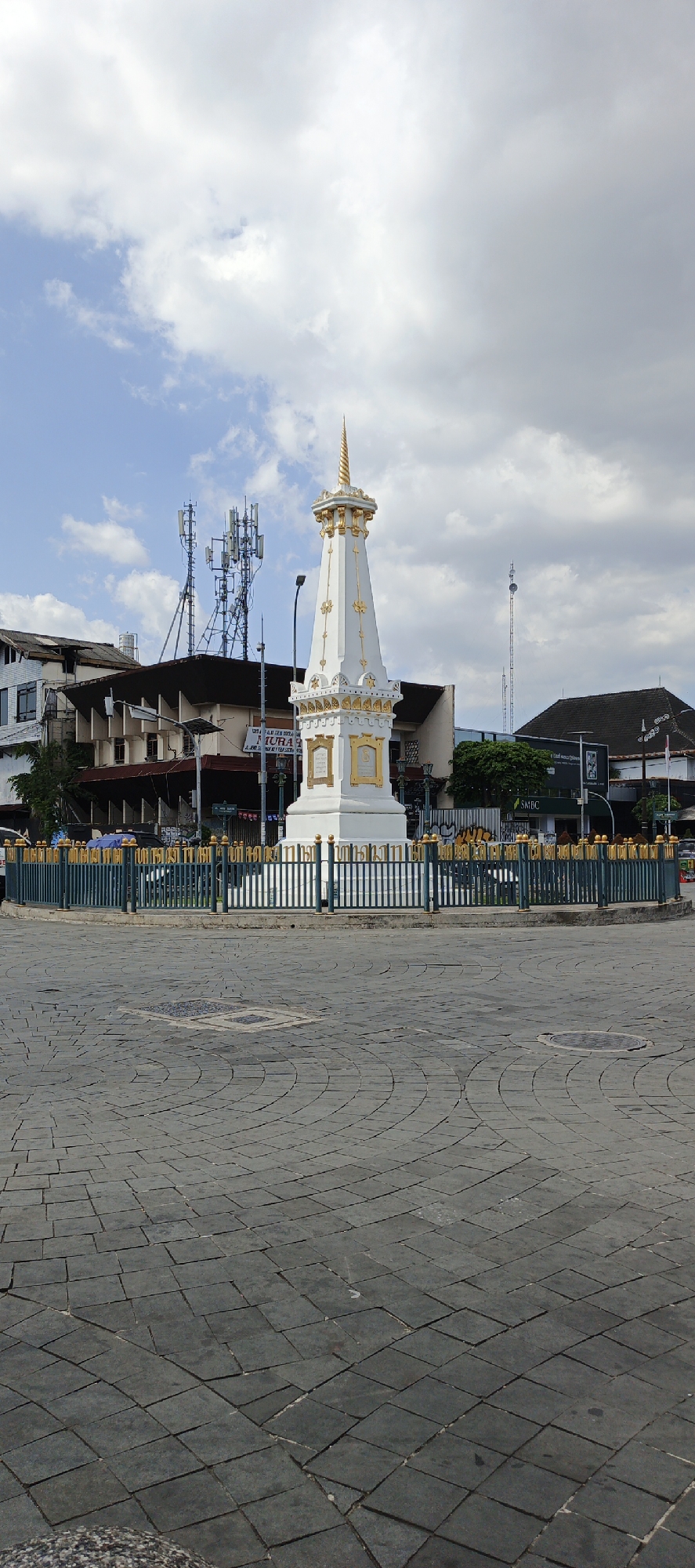 The image shows the iconic Tugu Yogyakarta, a monument located in the city of Yogyakarta, Indonesia. The monument is a tall, white obelisk with a golden spire, surrounded by a green and gold fence. The monument is situated in a circular plaza paved with dark gray square stones. In the background, there are several buildings of varying architectural styles, including one with multiple antennas on its roof. The sky is a bright blue with scattered white clouds, indicating a daytime scene. There are no people prominently visible in the image. The overall scene depicts a landmark in a city setting.
