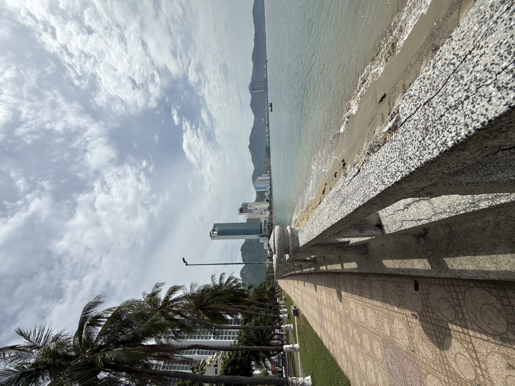 The image depicts a scenic view of the coast in Nha Trang, Vietnam. In the foreground, a paved walkway lines the shore, bordered by a concrete railing. Palm trees line the left side of the walkway, with their fronds reaching towards a cloudy sky. Beyond the walkway, the sandy beach meets the gentle waves of the ocean. In the distance, a line of buildings and lush green mountains form the backdrop, with a few boats visible on the water. The overall atmosphere is tranquil and idyllic, capturing the beauty of this coastal city.