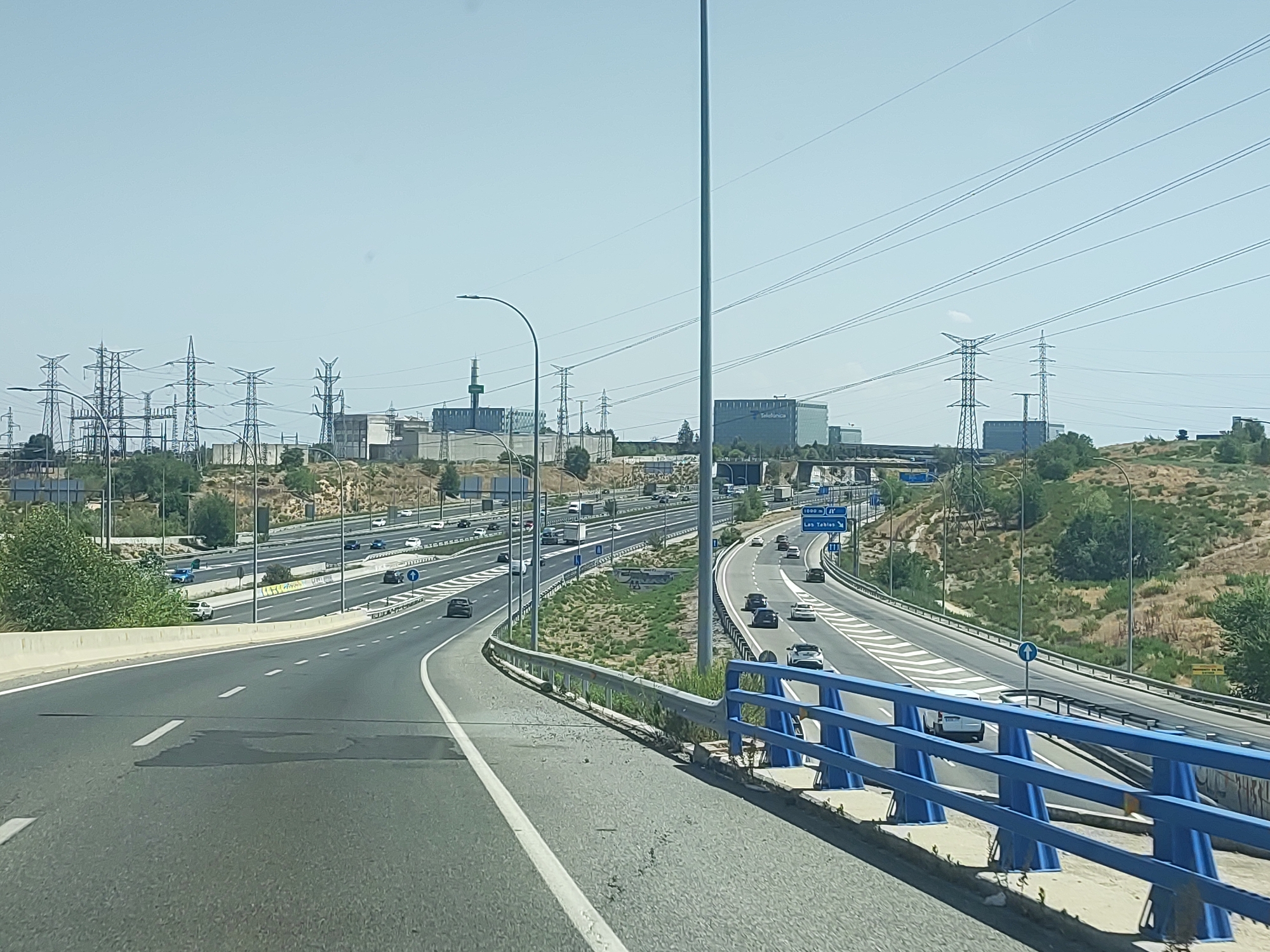 The image depicts a highway intersection in Madrid, Spain, likely in the northeastern part of the city. The highway is multi-laned, with a clear division between directions of travel. Several vehicles are visible on the highway, including cars and trucks. The environment is characterized by dry, grassy hillsides, sparse trees, and a bright, clear sky. Prominently featured in the background are numerous electrical pylons and power lines, suggesting the proximity of a significant electrical substation or power generation facility. In the distance, several large, modern buildings are visible, one of which appears to be a corporate headquarters, possibly with signage indicating "Telefónica." A blue metal guardrail lines the right side of the highway in the foreground. The overall scene suggests a sunny day in an urban or semi-urban landscape with substantial infrastructure.