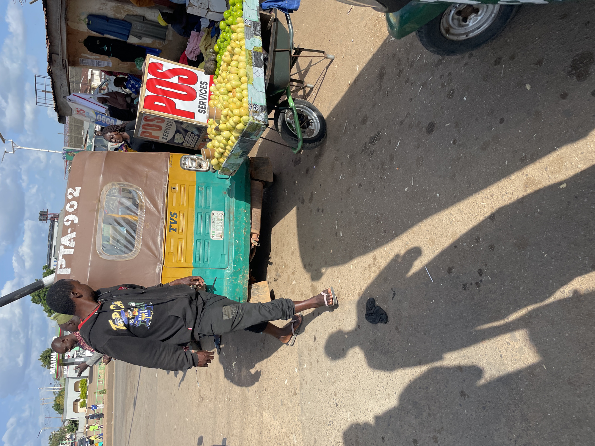 In Gura Top, Nigeria, a bustling outdoor market scene unfolds under a bright, cloudy sky. A man, wearing a black hooded sweatshirt with a colorful graphic and black pants, walks away from the camera. He is partially obscured by shadows cast by the sunlight. In the background, a yellow and green auto-rickshaw, identified by the license plate "PTA-902" and "TVS" branding, is parked. Adjacent to the auto-rickshaw, a cart is laden with a large pile of yellow fruits, likely lemons or limes, and green fruits. A prominent "POS SERVICES" sign, featuring red lettering on a white background, stands near the fruit cart. Several other individuals are visible in the background, some standing near the auto-rickshaw and others further down the street, engaged in market activities. The ground is paved and shows signs of wear, with scattered debris. The overall atmosphere suggests a typical day at a Nigerian roadside market.