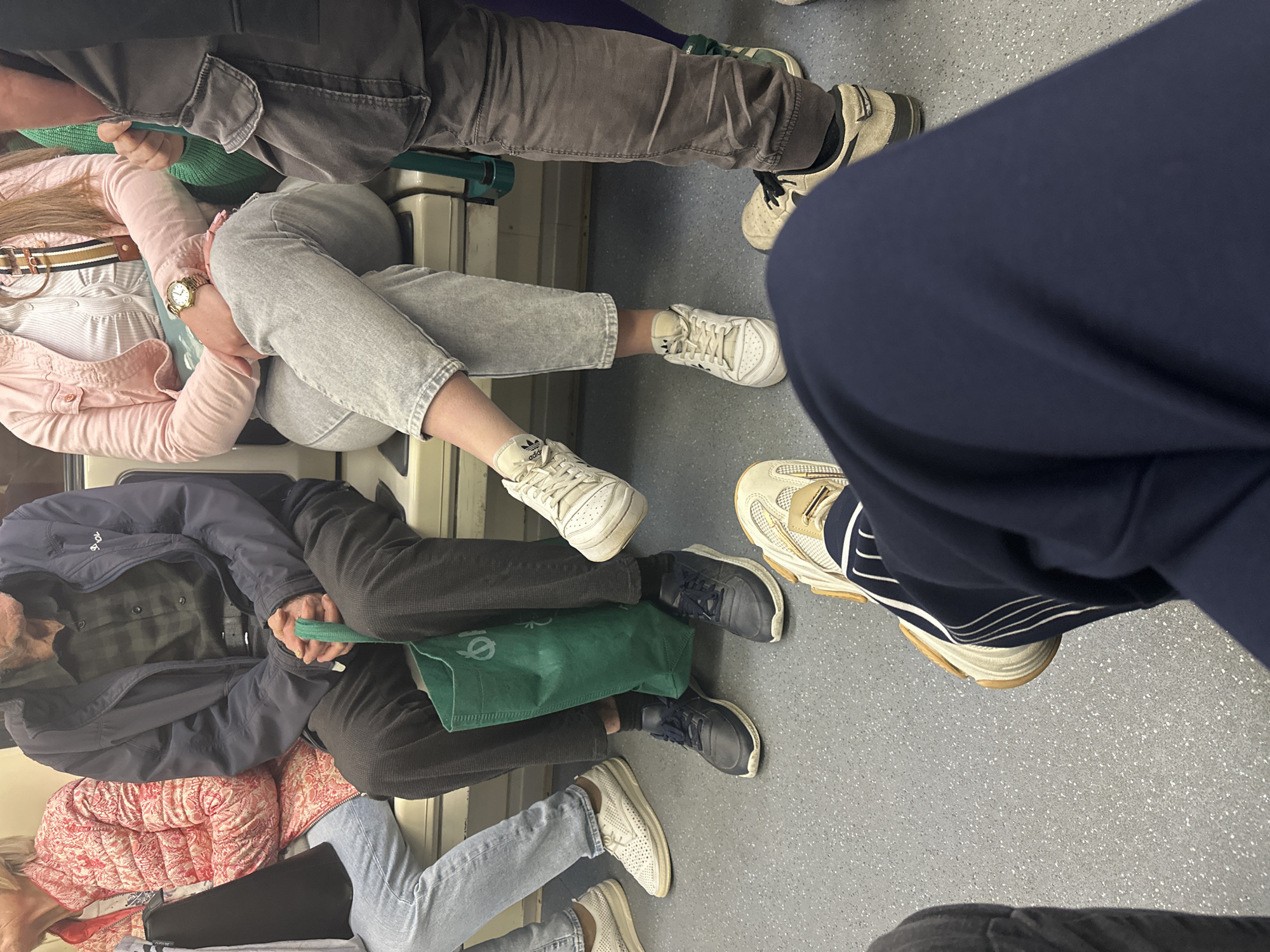 This is an eye-level indoor photograph showing the lower bodies and footwear of six individuals seated in what appears to be a subway car. The floor is gray.
Starting from the upper left corner, one individual is wearing gray pants and worn white sneakers. Next to them, another person is wearing light gray jeans and white sneakers with the Adidas logo. Further down, a person in a blue jacket is seen, sitting with a green tote bag resting between their legs, and wearing navy blue sneakers. Next to them, an individual with pink and red clothing is seen wearing light blue jeans and white sneakers. On the right, a person is wearing navy blue sweatpants and bulky, off-white sneakers. Partially visible is the footwear of a person with black pants.
No specific identifying information is provided about the location, indicating only a generic subway car setting. Therefore, the city of Sofia, Bulgaria cannot be confirmed.