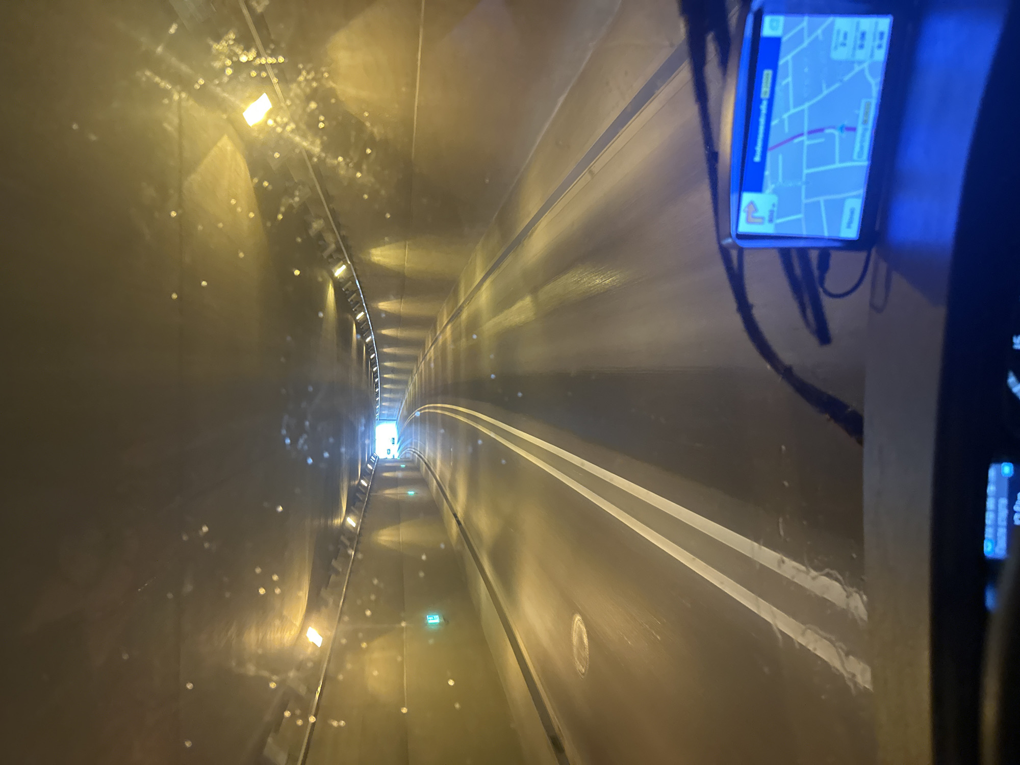 The image shows the interior of a vehicle driving through a tunnel in Memmingen, Germany. The tunnel walls are lit by yellow and white lights. There are green illuminated signs on the left wall. The road surface appears wet, with white lane markings visible. In the foreground, a GPS device is mounted on the dashboard, displaying a map. The windshield has streaks and smudges, indicating it may be wet or dirty. The perspective suggests the photo was taken from the driver's seat.