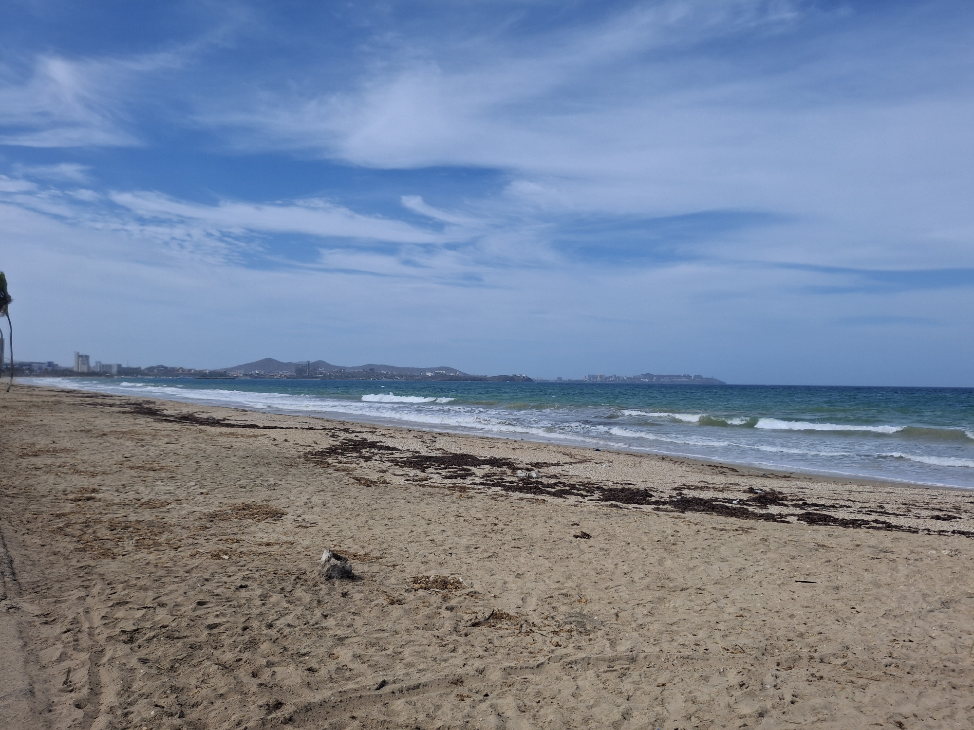 The image shows a beach scene in Porlamar, Venezuela. The sky is blue with scattered clouds. The ocean is a mix of blue and green, with small waves breaking on the shore. The beach is sandy and contains seaweed. In the background, buildings and hills are visible, indicating a coastal urban area. There are no people visible in the image.