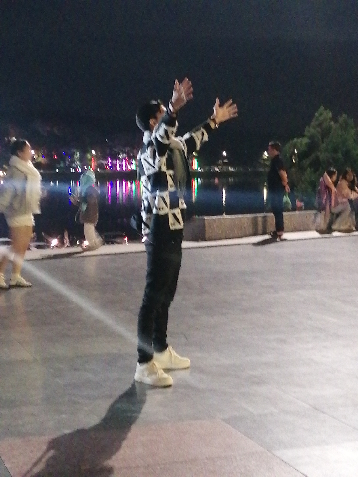 This image depicts a nighttime scene on a paved promenade, likely in Da Lat, Vietnam, with a body of water and a tree-lined background. In the foreground, a person is seen from the side, wearing a black and white checkered jacket, black pants, and white sneakers, with their arms raised upwards. Further back, to the left, another person is visible wearing a light-colored jacket and white shorts. Several other individuals are scattered in the background, near the water's edge, where colorful lights reflect on the surface. The promenade appears to be an urban area, possibly a park or a waterfront public space, given the presence of water, trees, and artificial lighting.