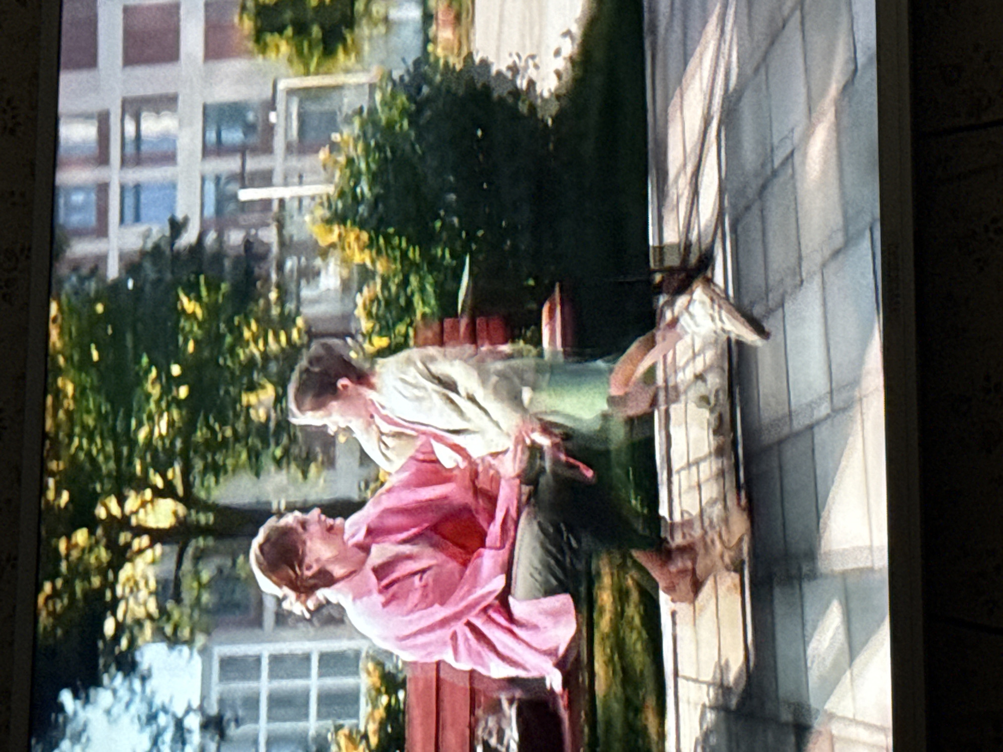 Two people are positioned outdoors on a paved area. A person in a pink top is carrying another person. A multi-story building is visible in the background, along with trees and a window. The scene is likely in Mytishchi, Russia.