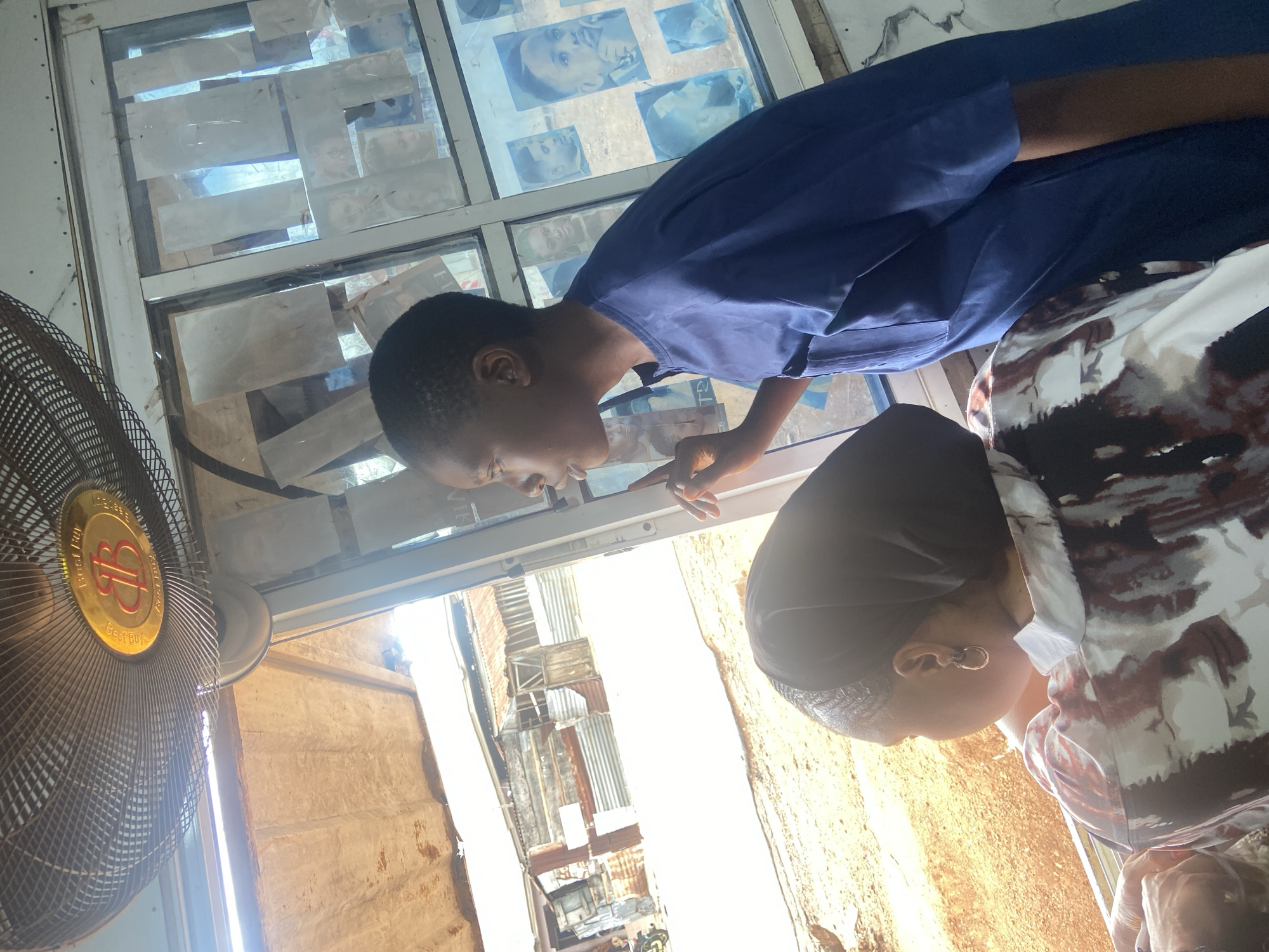 Two people, one wearing a blue shirt and the other wearing a patterned shirt with a black head covering, are looking at a window covered with pictures of faces, likely in a barber shop. A fan is visible in the foreground. The environment outside the window includes corrugated metal roofs and walls, indicative of a low-income area in Okengwe-Obehira, Nigeria. There is bright sunlight outside.