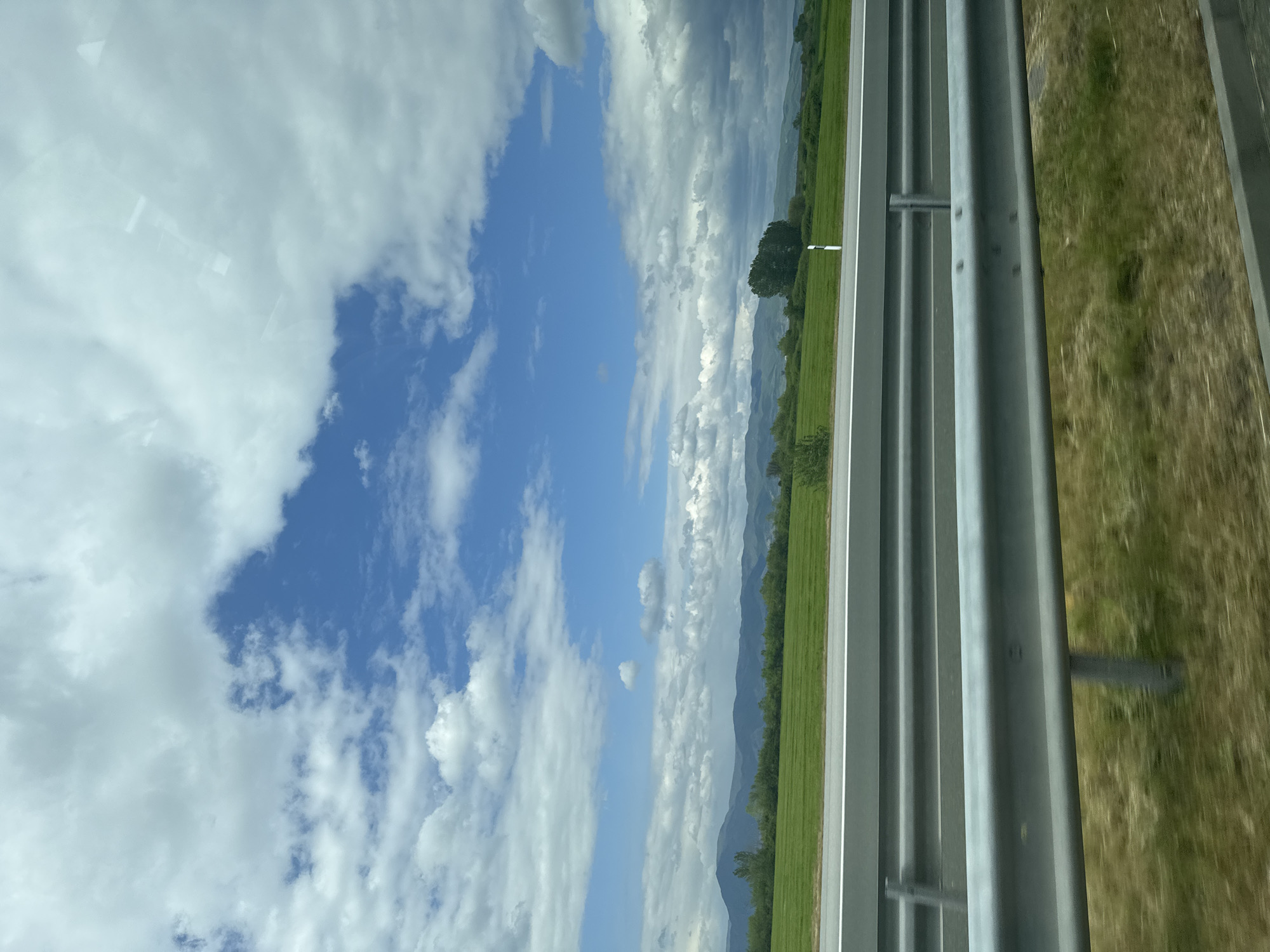 The image shows a view from a moving vehicle, likely a car, traveling on a road or highway. A metal guardrail runs along the right side of the frame. Beyond the guardrail is a grassy area. On the other side of the road, there is a field of green vegetation with a few trees. Further in the distance are mountains. The sky is blue with white clouds. The location is Zavoy, Bulgaria.