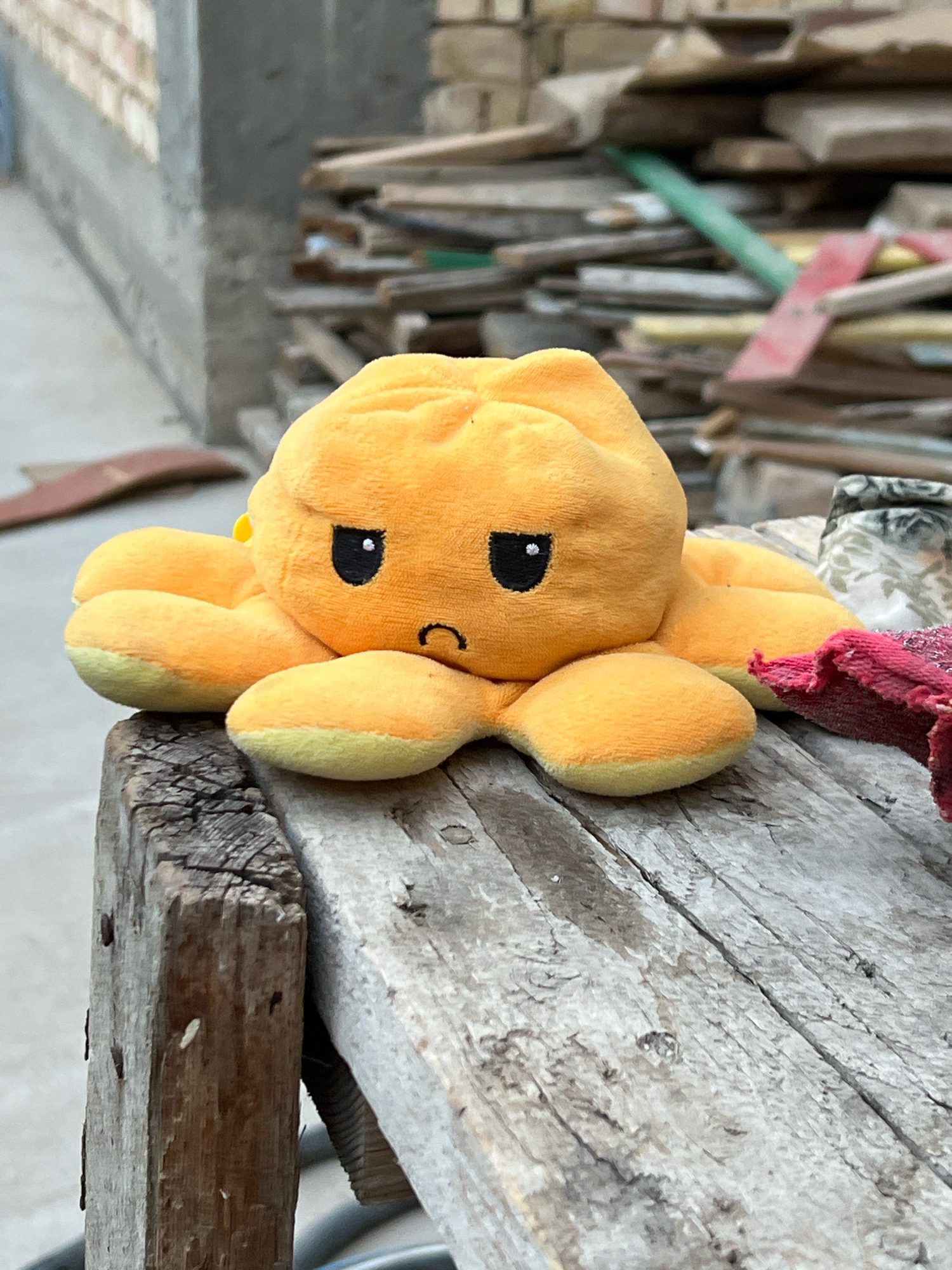 A plush, orange octopus toy with a sad expression sits on a weathered wooden surface. The toy has two black eyes and a downturned mouth. The background is out of focus and shows a brick wall, a pile of wooden planks, and some greenery. The lighting suggests it is daytime.