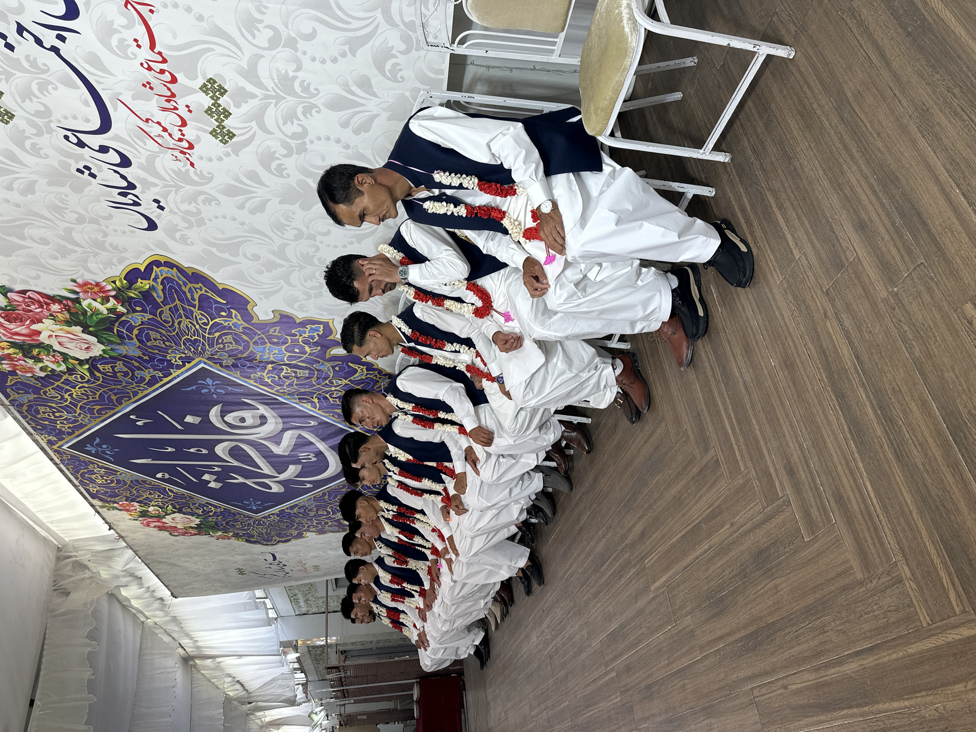 In a room with wooden parquet flooring in Quetta City Tehsil, Pakistan, a line of ten men sit on white metal chairs. The men wear white shalwar kameez, navy vests, and garlands of red and white flowers. The first man in line looks down and to the left. The backdrop features a sign with floral designs, geometric patterns, and Arabic calligraphy.