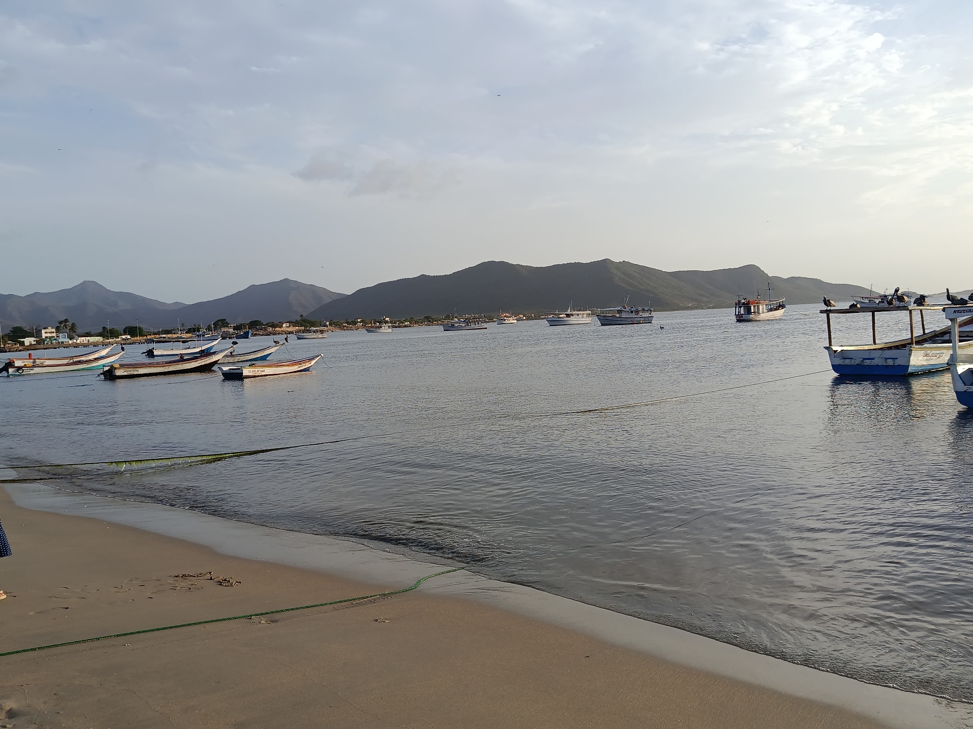 The image depicts a tranquil scene at the bay of Juan Griego, Venezuela. The foreground is a sandy beach with a few footprints and a green rope stretched across it. The water is calm and reflects the sky and distant mountains. Several small fishing boats are moored in the bay, some with pelicans perched on them. In the background, lush green mountains rise up, with a few buildings visible along the coastline. The sky is overcast with some hints of sunlight, suggesting it might be early morning or late afternoon. The overall atmosphere is serene and peaceful, characteristic of a small coastal town.