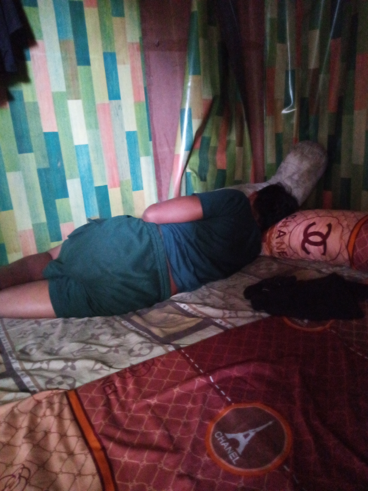 A person is lying on their side on a bed. The person is wearing a green shirt and green shorts. The bed has multiple patterned blankets. A pillow with a Chanel logo is next to the person. A dark shirt is also on the bed. The background consists of vertical striped curtains in various colors including green, blue, pink, and white. The scene appears to be indoors, possibly in a bedroom, in Candi Wates, Indonesia.