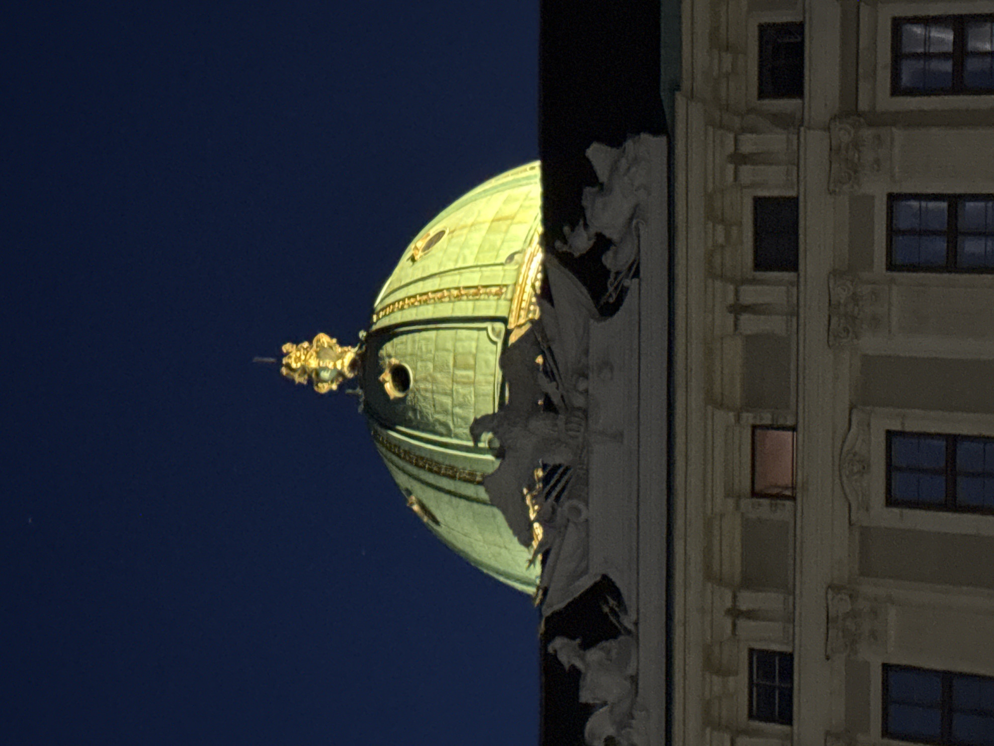 The image shows a close-up view of an ornate building facade and a green dome against a dark blue sky. The building on the right has multiple windows, some of which are illuminated from within with a warm, reddish glow, suggesting it is nighttime. The facade is decorated with classical architectural elements, including columns, moldings, and decorative sculptures of figures.
The dome on the left is a prominent feature, appearing to be made of a metallic material, possibly copper, with a green patina. It is illuminated by artificial lights, highlighting its rounded shape and architectural details. Golden decorative elements are visible on the dome and its supporting structure.
The overall scene suggests a grand, historical building, likely a church or a public institution, in Vienna, Austria. The time of day is clearly night or very late evening due to the dark sky and interior lighting. There are no visible people or text in the image. The composition is a vertical shot, with the building and dome tilted, possibly due to the camera's orientation or perspective.