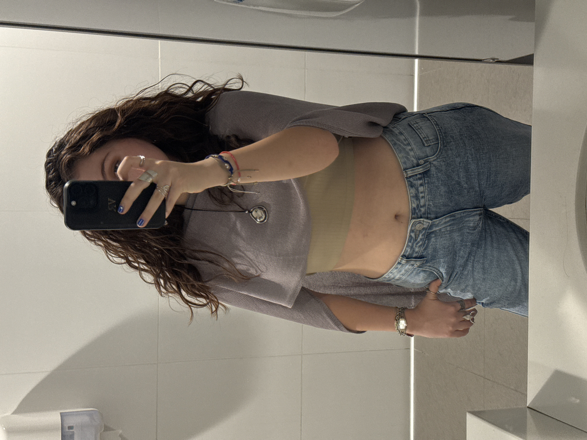 A young woman is taking a mirror selfie in what appears to be a public restroom. She is wearing a gray cardigan, a tan top that exposes her midriff, and light wash jeans. Her hair is long and wavy, and she has multiple rings on her fingers and bracelets on her wrist. She is holding a black smartphone to take the picture. The restroom has light gray tiles and a modern sink. There is no indication the location is in Mexico City, Mexico.