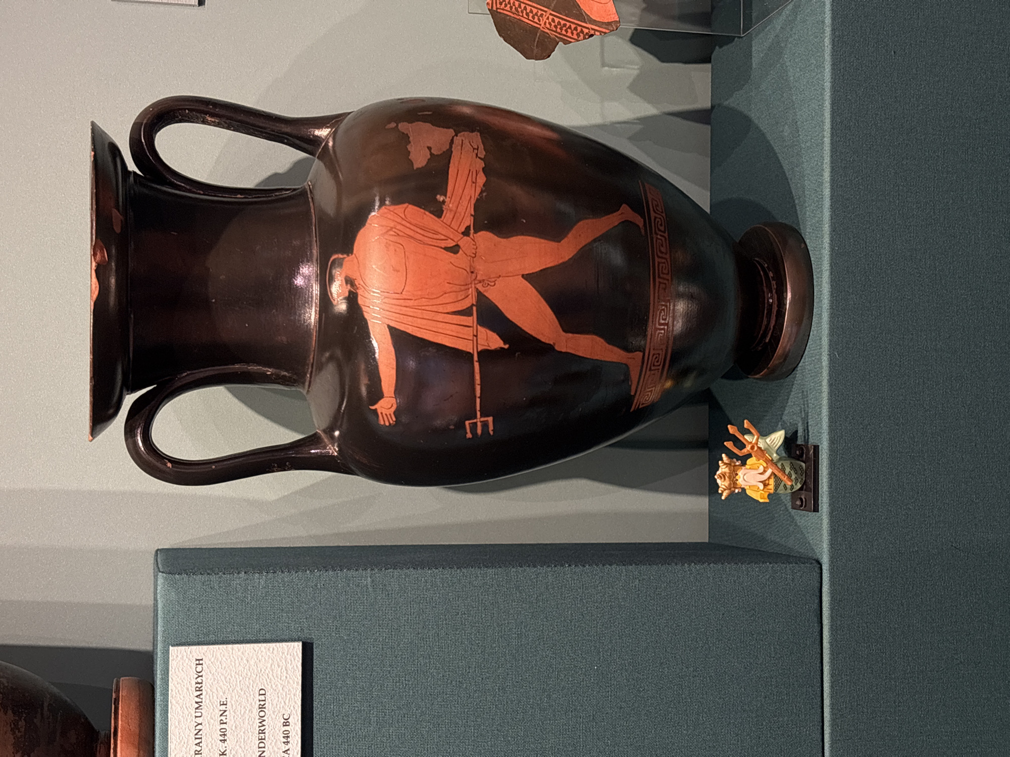 The image shows a black ceramic amphora, with a reddish-orange figure painted on its side. The figure is a male, clad in a draped garment, holding a trident. The vase is displayed in a museum setting, specifically in Krakow, Poland. There is a small decorative figure to the right of the vase. A label is partially visible in the lower-left corner.