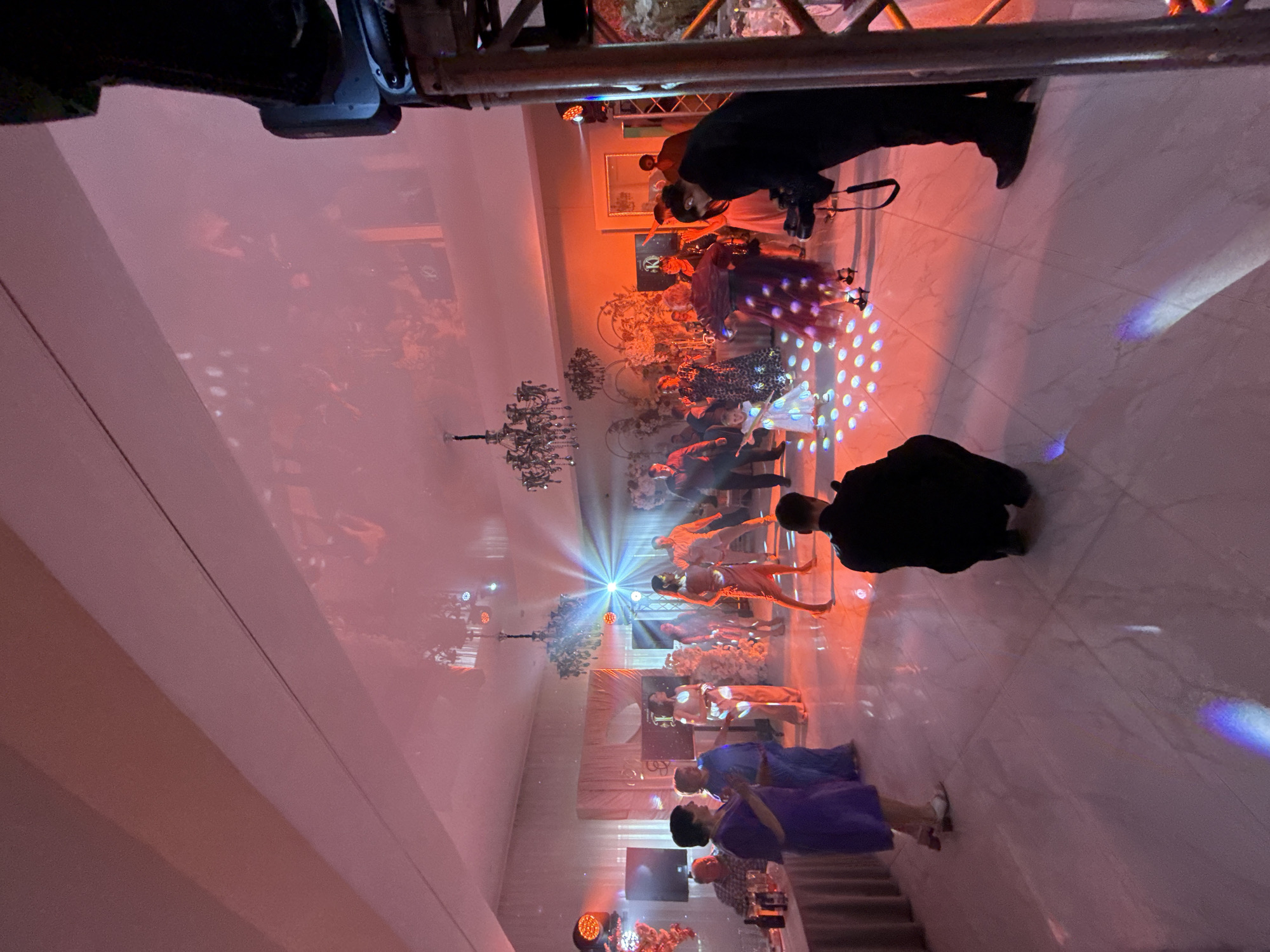 In a dimly lit banquet hall, guests are dancing under a pink and orange illuminated ceiling. A spotlight shines on the dance floor, creating a vibrant atmosphere. Several people are engaged in dancing, while others are conversing and observing. A photographer is crouched on the dance floor, capturing the event. The hall features decorative chandeliers and draped fabrics, adding to the elegant ambiance. There is no indication of this location being in the city of Perm, Russia.