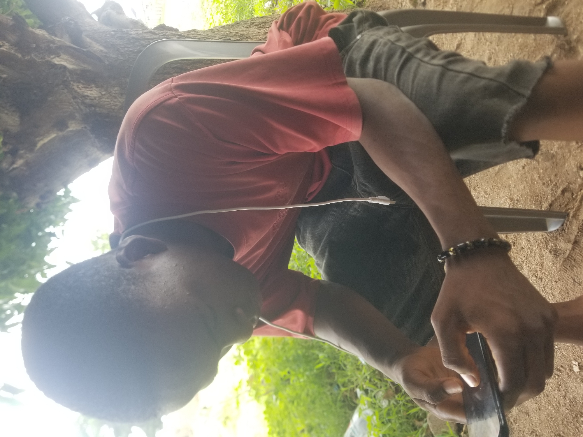 A person is seated on a plastic chair outdoors in Jauro Yinu, Nigeria. The person is wearing a red t-shirt and dark shorts, with a bracelet on their right wrist. A white cable is plugged into their phone, which they are holding and looking at. The scene is set against a backdrop of a tree and lush green vegetation, suggesting a natural, possibly rural environment. The lighting indicates it is daytime, with sunlight filtering through the trees. The person's focus on their phone suggests they are engaged in an activity such as browsing, texting, or using an app. No visible text is present in the image.