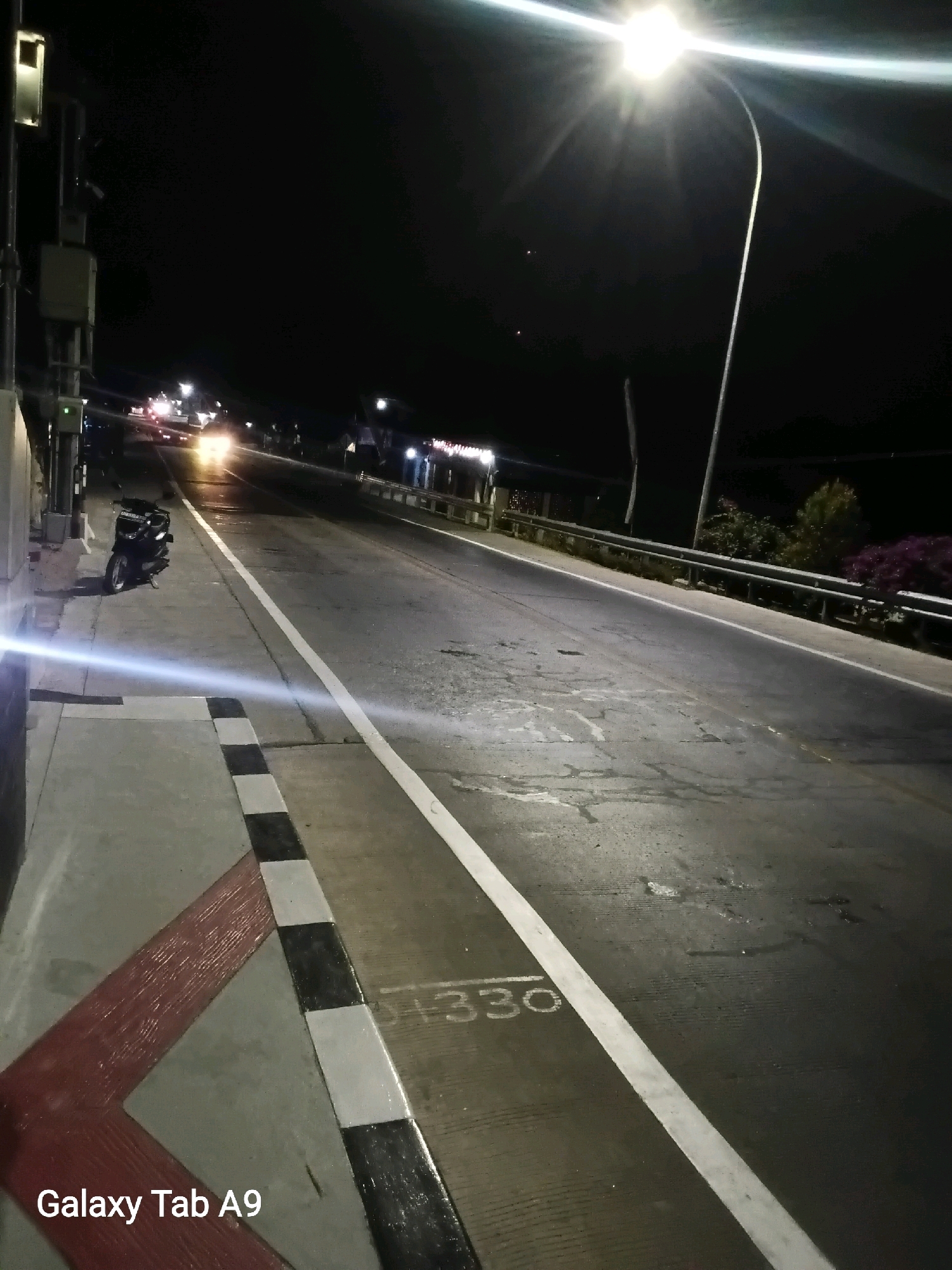 The image depicts a nighttime scene of a road in Bendo, Indonesia. A motorcycle is parked on the side of the road, which is illuminated by a bright streetlight and the headlights of approaching vehicles. The road appears to be paved with asphalt and has white lane markings. There is a sidewalk with black and white checkered markings. In the distance, there are buildings and vegetation. The sky is dark, indicating it is nighttime. The text "Galaxy Tab A9" is present in the lower left corner of the image, likely indicating the device used to capture the photo.