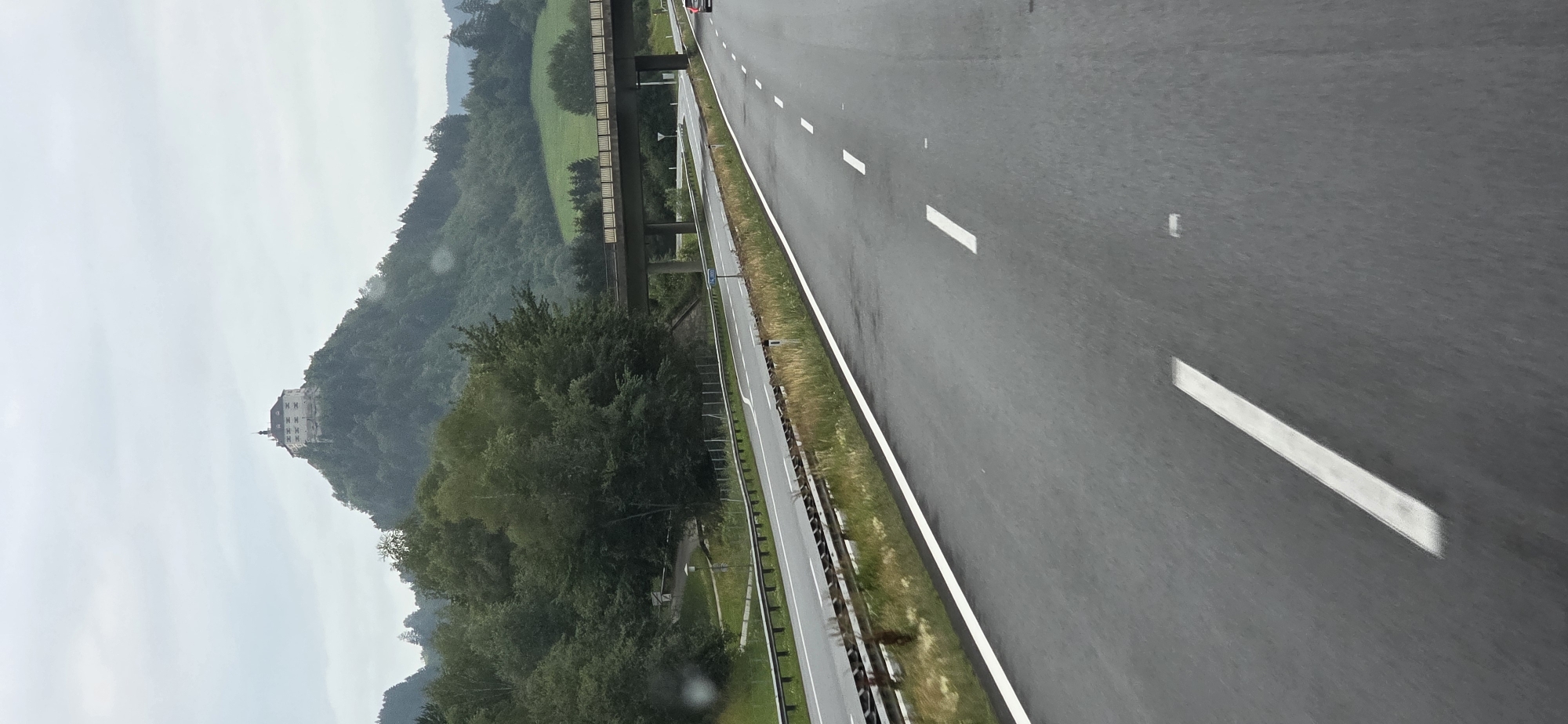 An overcast, grey day frames a view from a highway in Rottenmann, Austria. A large, multi-story building with a prominent tower sits atop a forested hill in the distance, likely a castle or similar historic structure. A multi-lane highway stretches into the distance, with wet asphalt indicating recent rain. White lane markings are clearly visible on the road. A pedestrian overpass crosses the highway in the middle ground, supported by concrete pillars. Green trees and rolling hills are visible on either side of the highway, with some patches of green grass on the hills. A single red car is visible on the highway in the distance, heading away from the viewer. The scene suggests a travel or transit moment, capturing the unique landscape of the Austrian region.