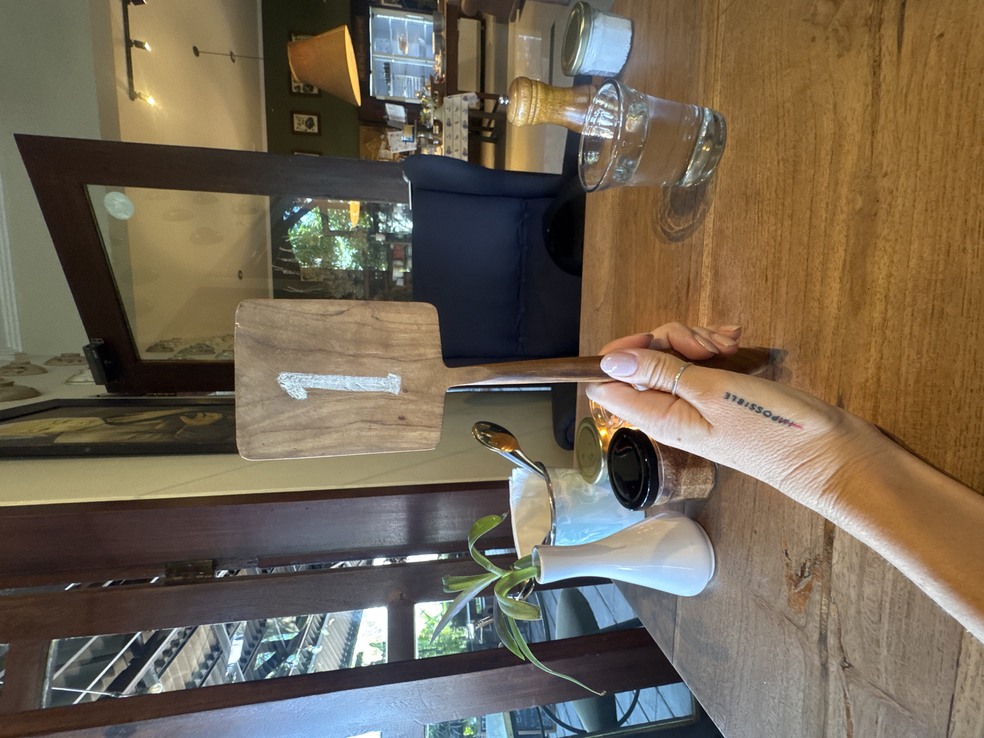 The image depicts a table setting in what appears to be a cafe or restaurant. A woman's arm rests on the wooden table, bearing a tattoo of the word "Impossible" with a line through it, making it read "I'm Possible." On the table are a wooden number stand marked with the number "1," a small white vase with greenery, a condiment holder, and a glass jar with a wooden top. There is also a salt shaker. The view through the window suggests an outdoor or semi-outdoor setting, typical of the North Kuta, Indonesia region. A dark blue upholstered chair is partially visible in the background. The scene is brightly lit with natural light.