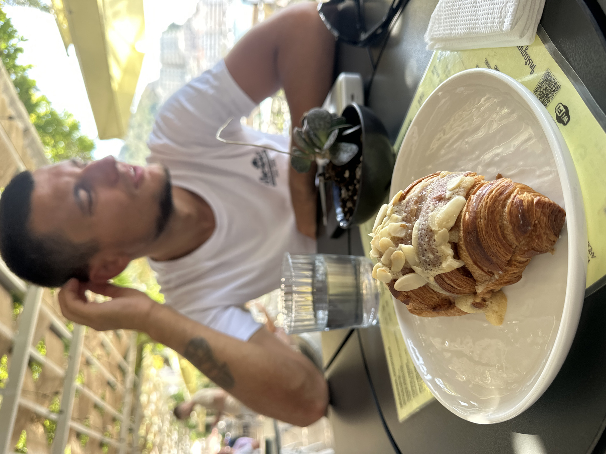 The image shows a close-up of a croissant topped with cream and almonds on a white plate, situated on a black table. Behind the croissant, a glass of water, a small potted plant, and sunglasses are visible. A man, partially in focus, is seated at the table, touching his ear. He is wearing a white t-shirt. The background suggests an outdoor setting, possibly a cafe or restaurant, with a covered structure and foliage overhead, indicative of a warm climate. The scene is likely located in Rafailovići, Montenegro, based on the surrounding architecture and vegetation that are common in coastal Mediterranean towns.