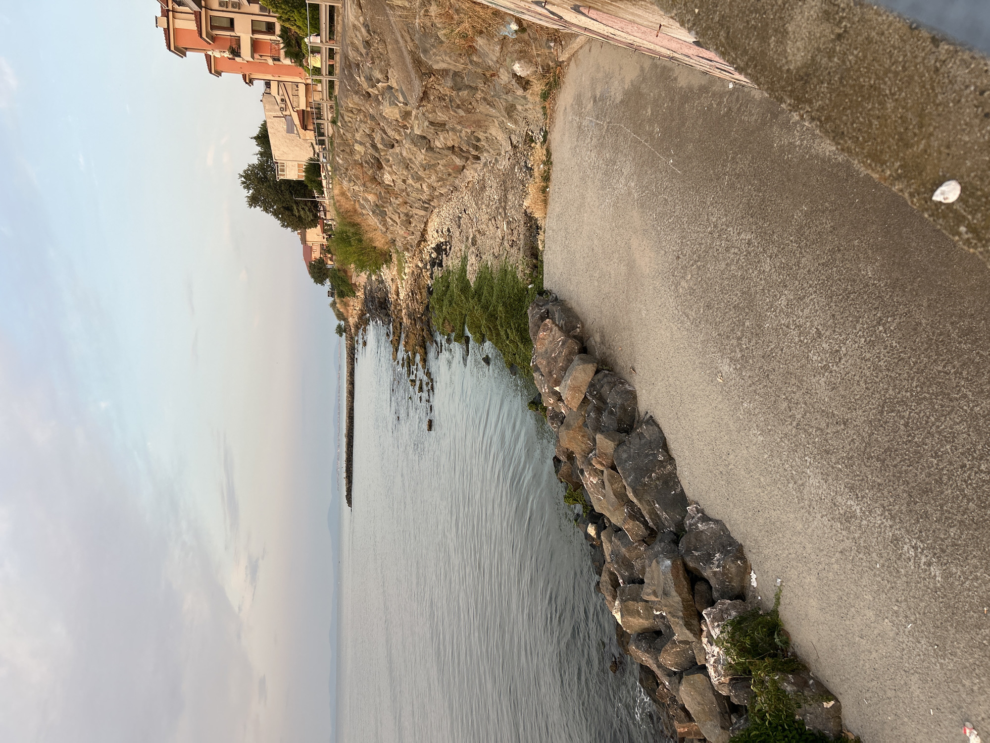 The image depicts a coastal scene in Ravda, Bulgaria, featuring a rocky shoreline and buildings. In the foreground, a concrete path slopes down towards the water, bordered by large, dark rocks. Some green vegetation grows among the rocks on the left. The sea is calm and grey, with a distant landmass visible on the horizon. To the right of the path, a stone embankment supports a grassy area and then a row of buildings. The buildings are multi-story structures with orange and pink facades, typical of coastal resorts. A wooden fence runs along the embankment in front of the buildings. The sky is partly cloudy with a soft, diffused light, suggesting either early morning or late afternoon. There are no visible people or animals in the image.