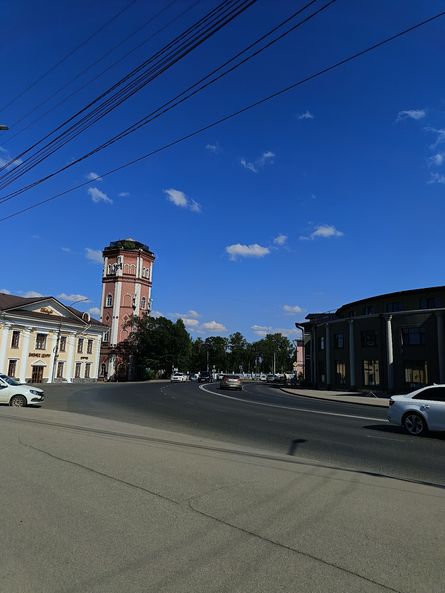 A street scene in a city is captured under a clear blue sky with scattered white clouds. In the mid-ground, a red and white tower with multiple windows stands prominently. To the left of the tower is a beige building with columns and arched windows, labeled with Cyrillic text. To the right of the tower, a dark, modern building with large windows and columns curves away from the viewer. Several cars are on the road, with one white car in the foreground on the left and another white car further down the road on the right. The road surface is asphalt, showing some cracks. Power lines are visible stretching across the top of the frame against the sky. Trees line the background between the buildings.