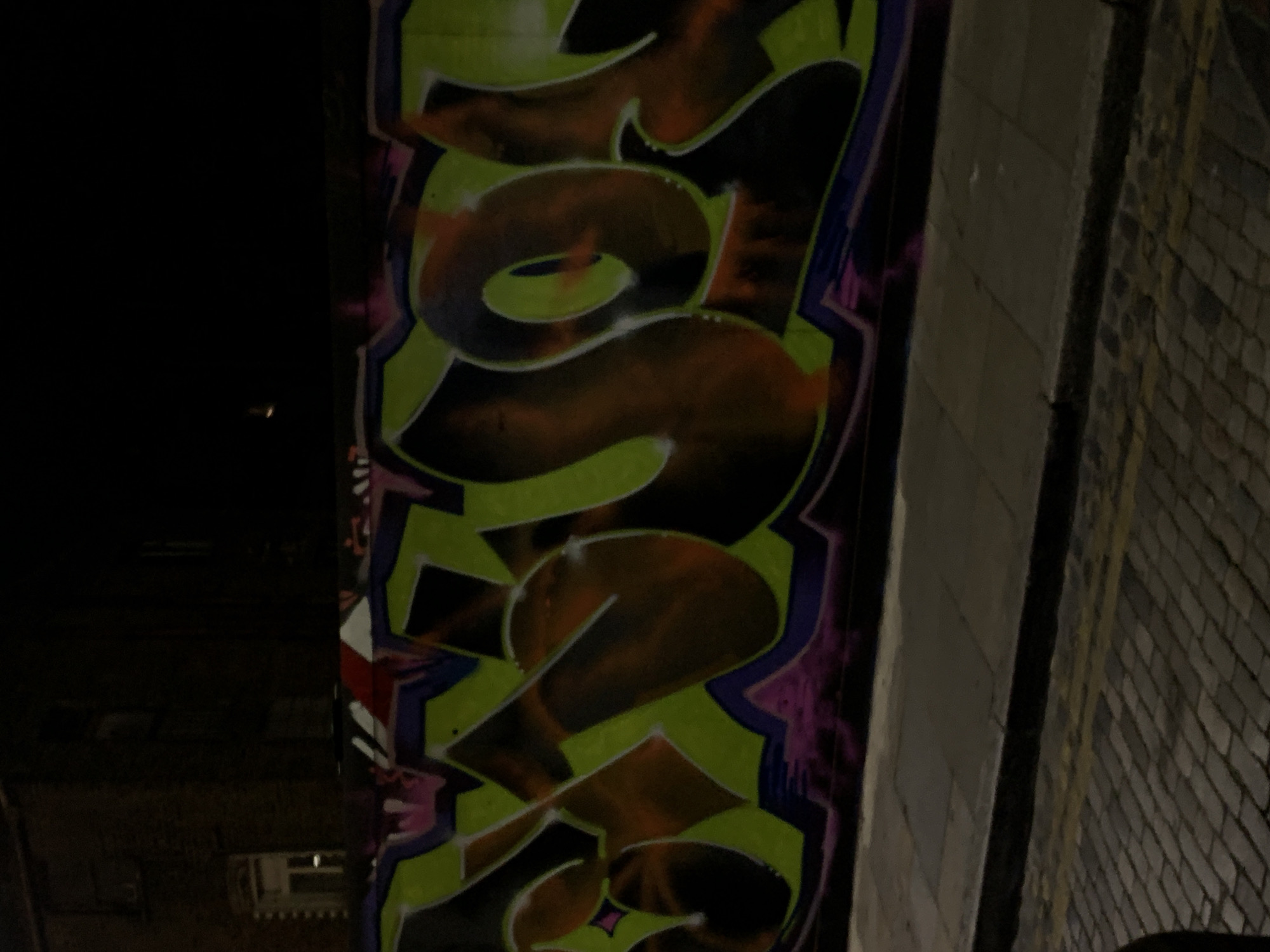 The image captures a vibrant, multi-colored graffiti artwork on a dark wall, likely part of an urban structure such as an underpass or bridge support. The scene is illuminated by artificial light, suggesting it is nighttime or a very dimly lit covered area.
The central subject is a large, stylized word rendered in graffiti script, predominantly featuring bright lime green outlines and a dark orange-brown fill. Purple and hints of pink and white are used for additional detailing and outlines, creating a bold, three-dimensional effect. The decipherable text on the graffiti spells out "SOUTH".
To the left of the graffiti, the background is completely dark, obscuring any details. To the right, the wall continues, transitioning into lighter concrete blocks and then into a textured brick wall with a diagonal pattern, possibly a metal fence or mesh overlaying it. A dark vertical pipe or beam runs along the junction of the graffiti wall and the brickwork.
No people or direct activity are visible. The atmosphere is starkly urban and nocturnal. The blend of vibrant street art against the grim, industrial backdrop is characteristic of hidden city spaces. Given the location context, this type of street art is common in London's underpasses and railway arches.