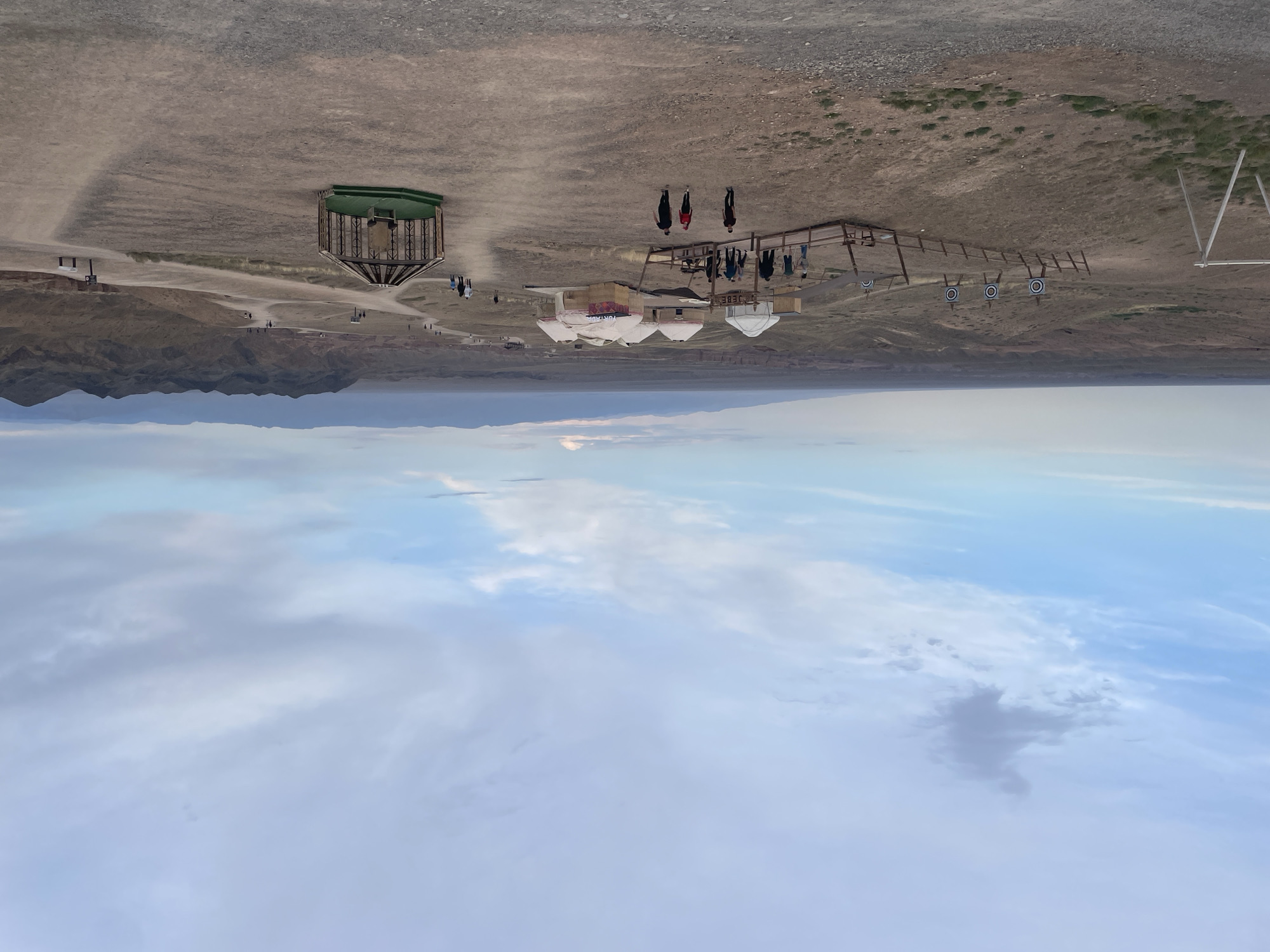The image depicts a panoramic view of a tourist encampment in a desert landscape, likely the Charyn Canyon area near Tamerlik, Kazakhstan. Several yurts, traditional nomadic dwellings, are clustered together. In the foreground, a group of people are walking on a dirt path. To the left, an archery range with targets is visible. Further into the distance, rolling hills and mountains form the horizon under a cloudy sky. A unique, gazebo-like structure with a green platform stands on the right side of the image. The overall scene suggests an outdoor recreational area catering to tourists exploring the natural beauty of the region.