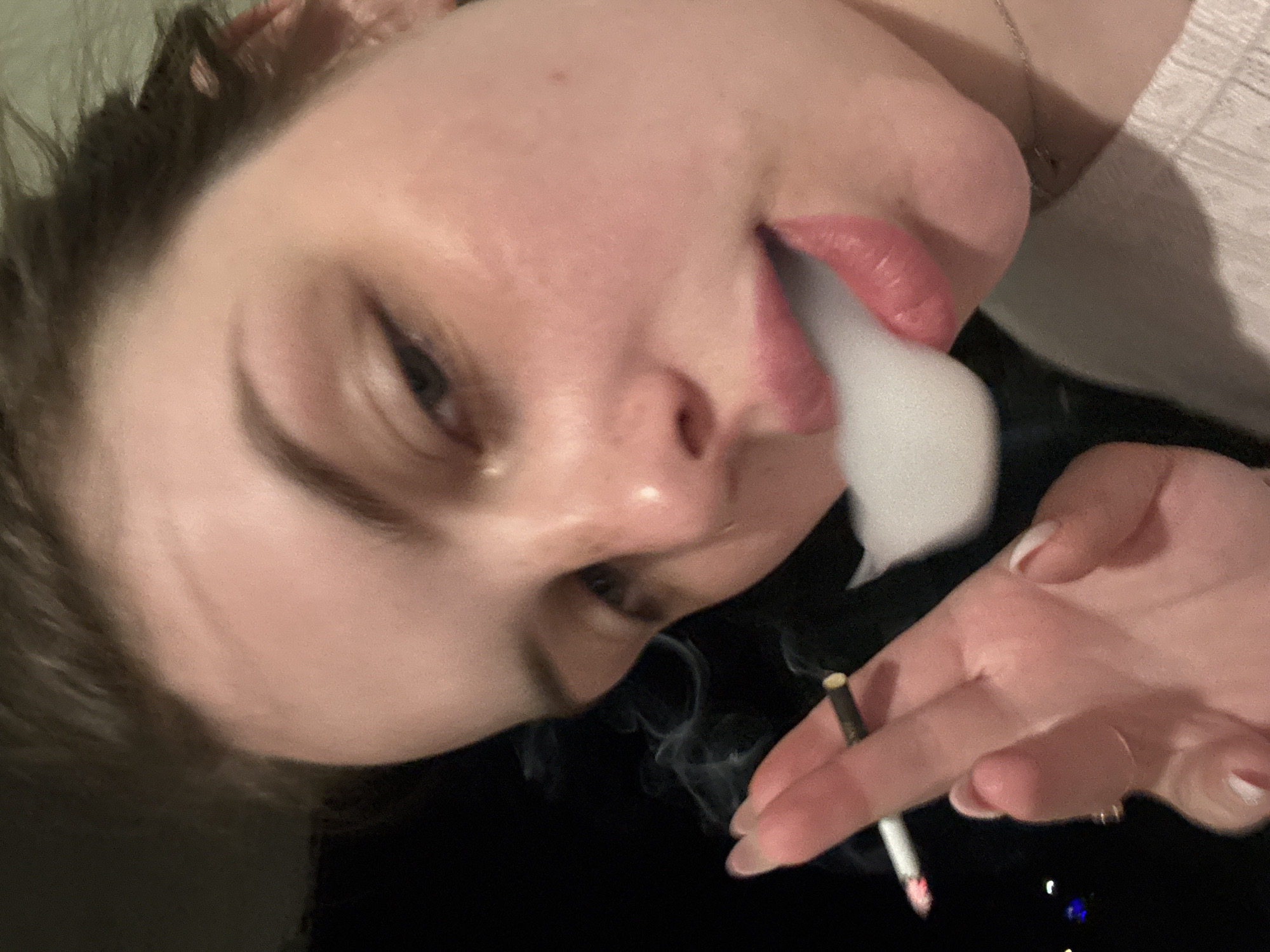 A close-up, angled portrait captures a young woman smoking. Her face, tilted and looking downwards, features fair skin and dark hair partially visible at the top and left. Her full lips are slightly parted, exhaling a prominent plume of white smoke. In her right hand, a lit cigarette is held between her fingers, emitting faint tendrils of smoke; its tip shows a glowing ash. Her fingernails are manicured with light-colored polish. A thin necklace chain is visible around her neck, and she wears layered clothing, with a light-colored garment over a darker one. The background is dark and out of focus, containing indistinct glimmers of light, suggesting a nighttime or dimly lit indoor setting. The woman's expression is neutral, concentrated on the act of exhaling smoke. There is no visible text. The scene depicts a casual moment in Chornomorskoe, Ukraine.