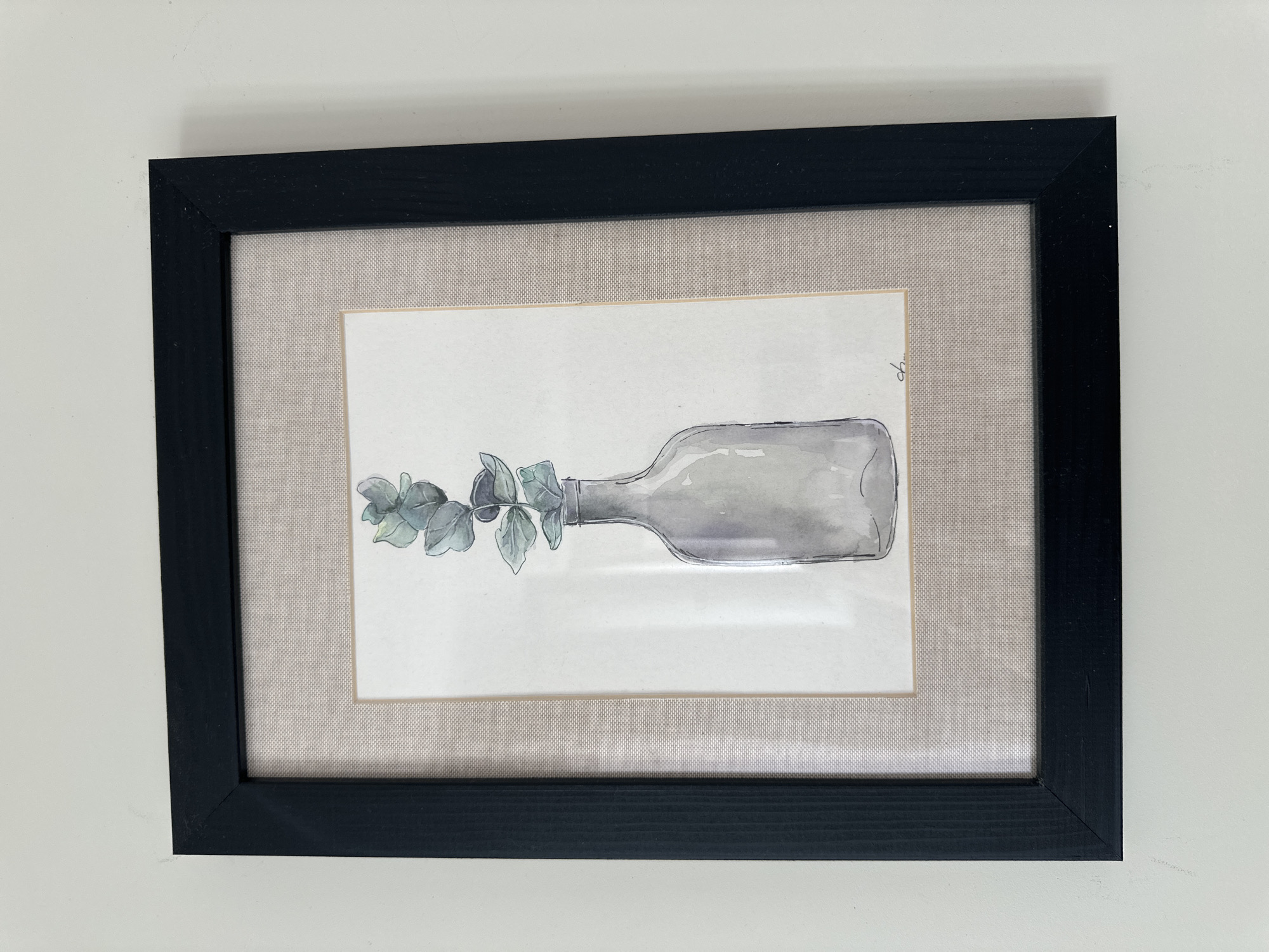 A framed watercolor painting of a glass bottle with eucalyptus stems is displayed on a white wall. The painting is in landscape orientation and is matted with a textured beige fabric and a thin cream-colored mat. The frame is black and has a slightly distressed finish. The painting depicts a clear glass bottle with a narrow neck, filled with several sprigs of eucalyptus leaves. The leaves are rendered in shades of green and blue-grey. A small, dark signature is present in the bottom right corner of the painting. The wall appears to be painted a light, neutral color. There are no people or other objects visible in the immediate vicinity of the framed artwork. The scene is indoors. This image is not specific to Minsk, Belarus, as there are no identifying features or landmarks present.