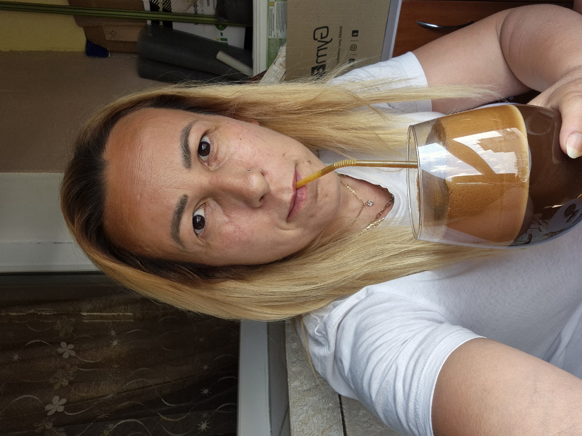 A woman with blonde hair and a white t-shirt is drinking from a glass of coffee with a straw. She is holding the glass with her right hand and has a straw in her mouth. The background is blurred but appears to be indoors with some boxes stacked up. The lighting suggests it is daytime. The woman has a neutral expression on her face. There is text visible on a box in the background, which appears to be in Cyrillic script.