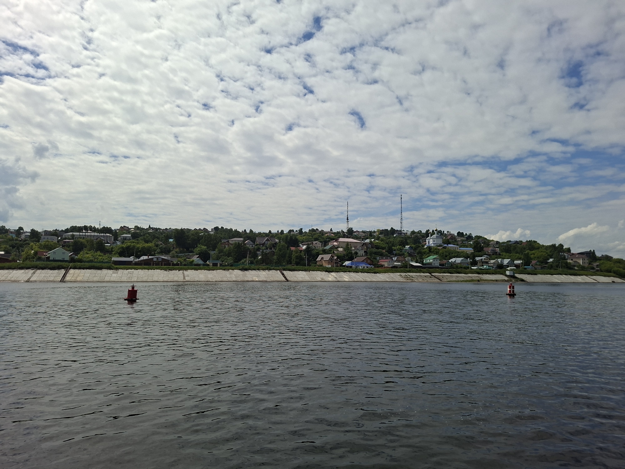 A wide shot captures a body of water with a distant, tree-lined riverbank dotted with houses and buildings. The sky is filled with a dense, patchy cloud cover, with glimpses of blue sky peeking through. Two buoys, one red and one red and white, are visible in the water. The riverbank is lined with a concrete embankment. The overall scene depicts a tranquil, rural or small-town setting.