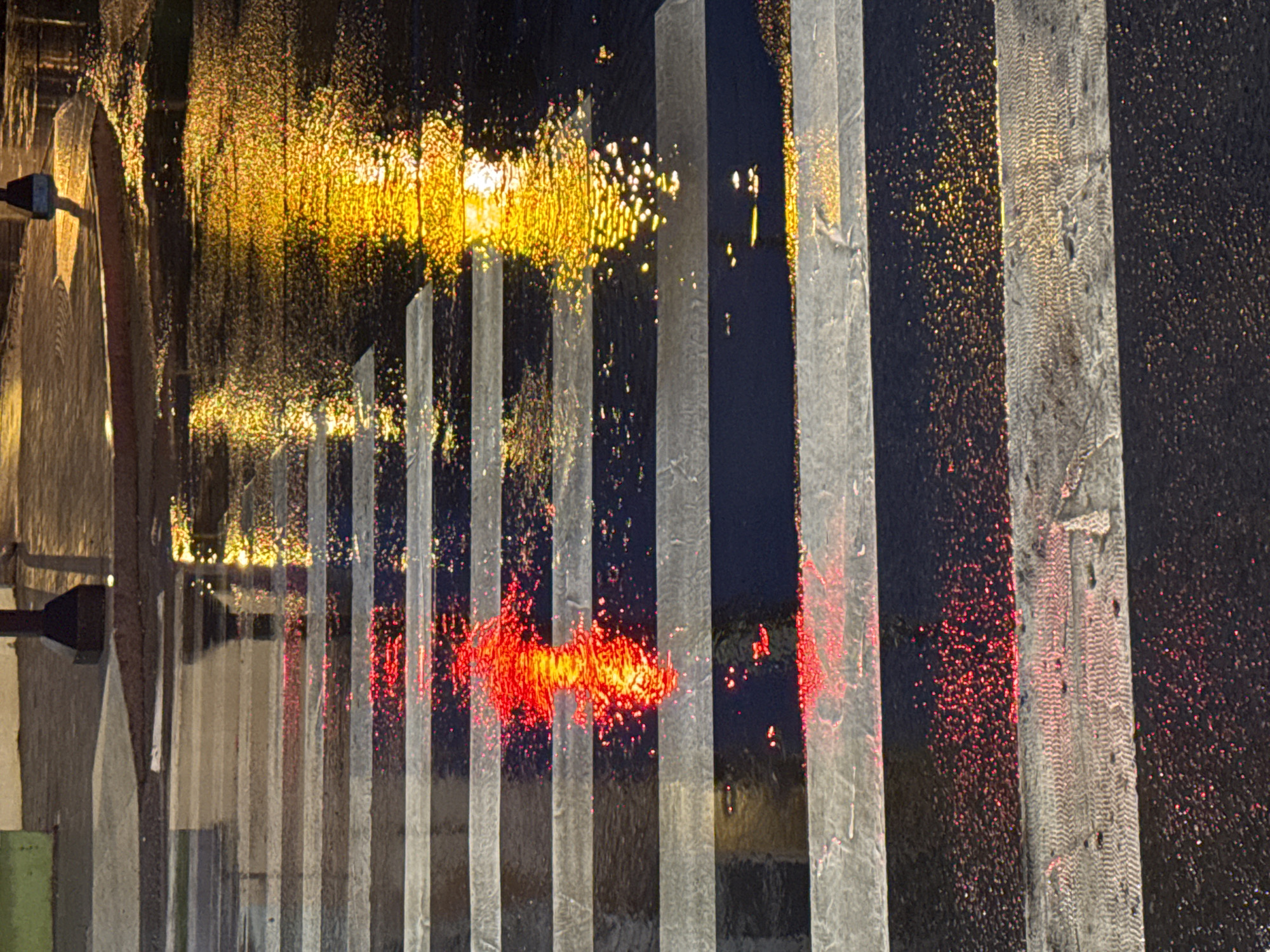 The image shows a wet crosswalk at night, with reflections of red and yellow lights on the asphalt. The crosswalk stripes are white and black. The reflections are distorted by the water, creating abstract patterns of light. The scene appears to be in a city, with a lamppost visible in the background. The overall mood is somewhat moody and atmospheric due to the low light and wet conditions. The image does not provide any specific information about the city of Krasnoyarsk, Russia, or any people or actions occurring in the scene.