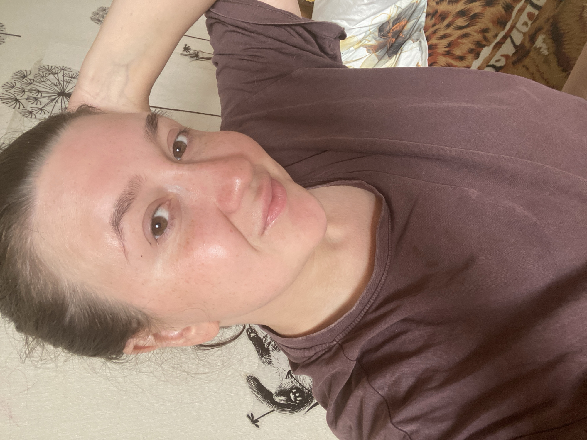 A young woman is captured in a close-up selfie, her face turned slightly towards the camera with her right arm resting behind her head. She appears to be indoors, in what looks like a bedroom. The background shows a wall with a patterned wallpaper featuring dandelion silhouettes and some animal-like drawings. To the right of the woman, a bed is visible with patterned bedding. She is wearing a plain, dark purple t-shirt. Her hair is pulled back loosely, and she has a natural, makeup-free appearance, with freckles visible on her cheeks. The lighting suggests an indoor setting, possibly with natural light coming from the side. There is no indication of the city of Chaikovsky, Russia, in the image or its content.