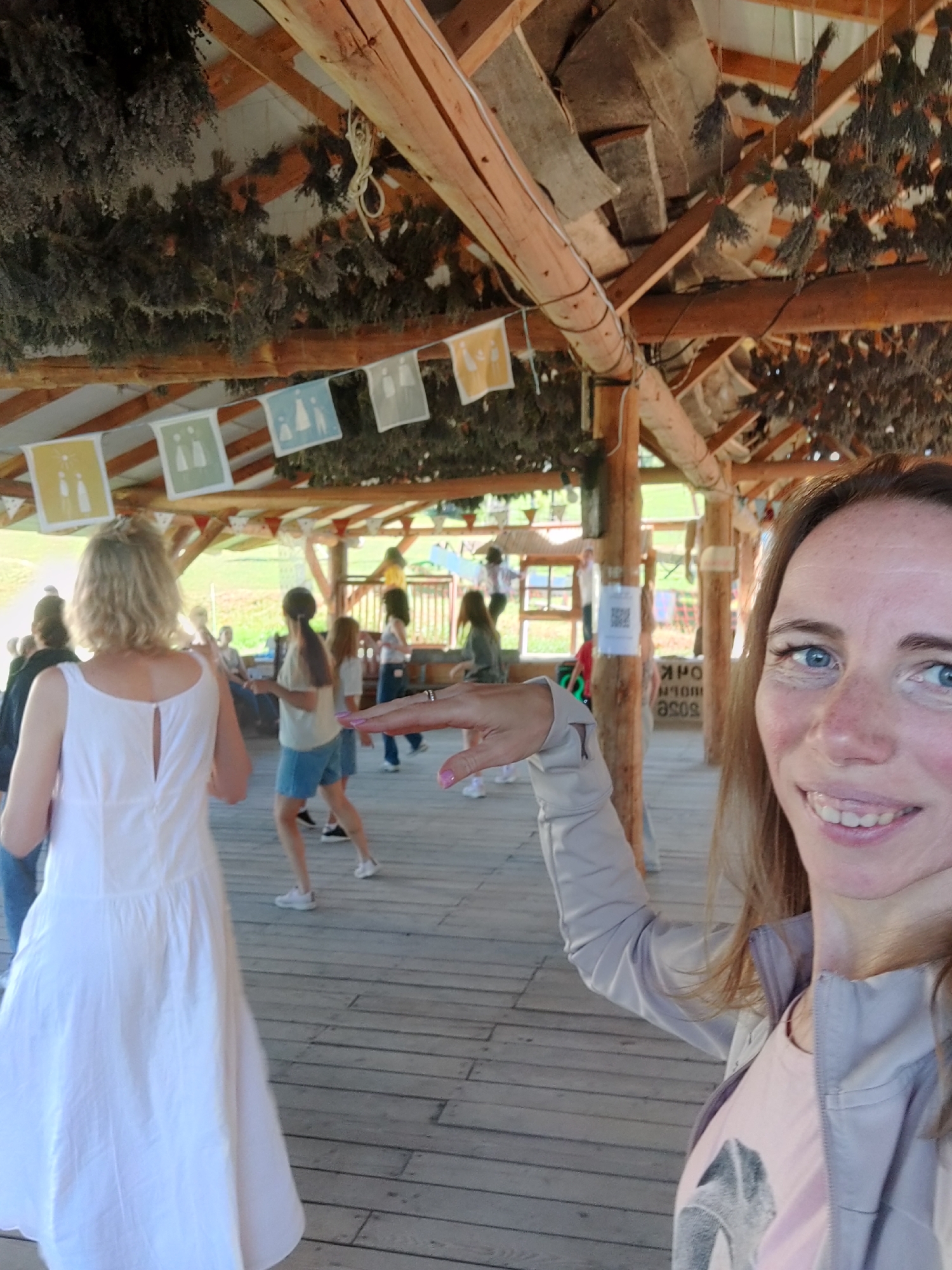 In Izky, Ukraine, this image captures a gathering under a rustic wooden pavilion, possibly at an outdoor event. The pavilion's roof is supported by thick wooden beams, with dried bundles of herbs or flowers hanging from the structure. A string of colorful fabric banners featuring simple white icons is strung across the ceiling. The floor is made of weathered wooden planks.
In the foreground, a woman with light brown hair and blue eyes is smiling and sticking out her tongue, her face close to the camera. She is wearing a light-colored jacket over a pink t-shirt with a graphic print. Her right hand is extended towards the center of the frame, with a ring on her finger.
In the midground, a woman with blonde hair, wearing a white dress, is seen from behind, facing away from the camera. Several other people are scattered throughout the pavilion, some appearing to be dancing or moving. One woman in the mid-left is wearing denim shorts and a light-colored top. Another woman in the mid-right is wearing a green top and dark pants.
In the background, glimpses of a green, outdoor landscape are visible through the open sides of the pavilion. A white sign with a QR code is attached to a wooden post. The overall atmosphere suggests a casual, communal event.