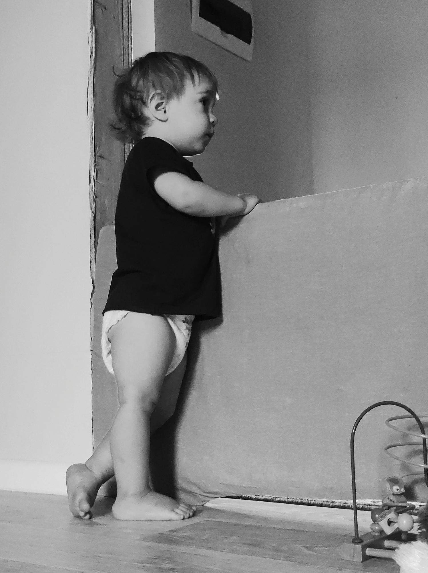 A black and white, eye-level photograph depicts a toddler standing upright in an indoor environment, utilizing a low barrier for support. The scene is located in the city of Фоки, Russia. The child is observed in profile, facing right, wearing a dark short-sleeved t-shirt and a light-colored diaper. Their light-toned, short, wavy hair is visible. Both hands are positioned on the upper edge of a light-colored, rectangular surface, which serves as a support. The child's bare feet rest on a light-colored floor, with the left heel slightly elevated. The gaze is directed forward and slightly downward. To the immediate left, a textured wall or door frame exhibits signs of surface degradation. The background to the right consists of a plain wall, with a rectangular feature, possibly a vent or upper window frame, visible near the top. In the lower right foreground, a portion of a toy bead maze is visible, consisting of a dark arched wire, several spherical beads, and a small animal-like figure.