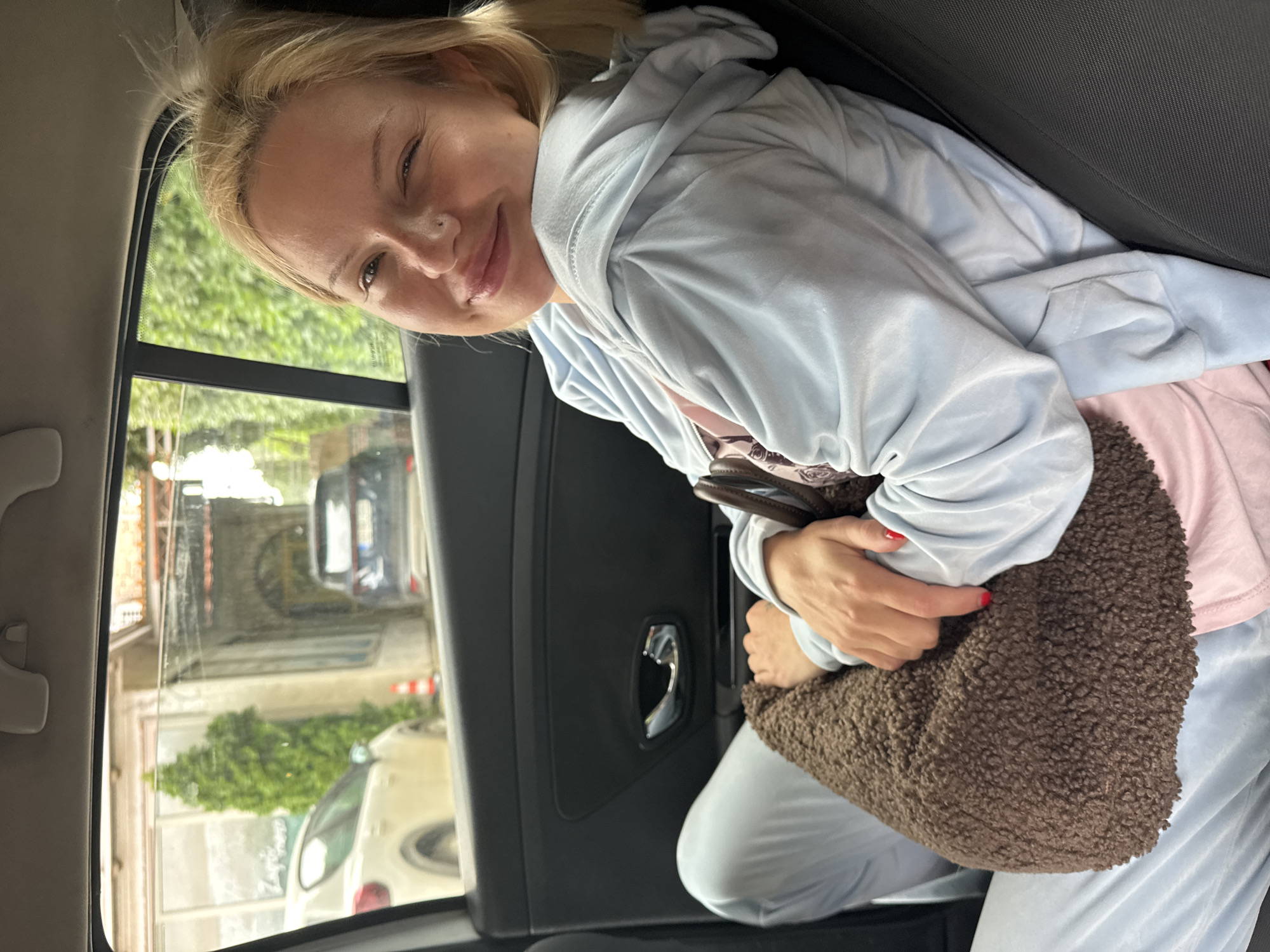 A woman with blond hair and a light blue hoodie is sitting in the back seat of a car. She is holding a brown, furry bag. The car window is open, revealing a street scene in Budva, Montenegro, with buildings and other cars visible.