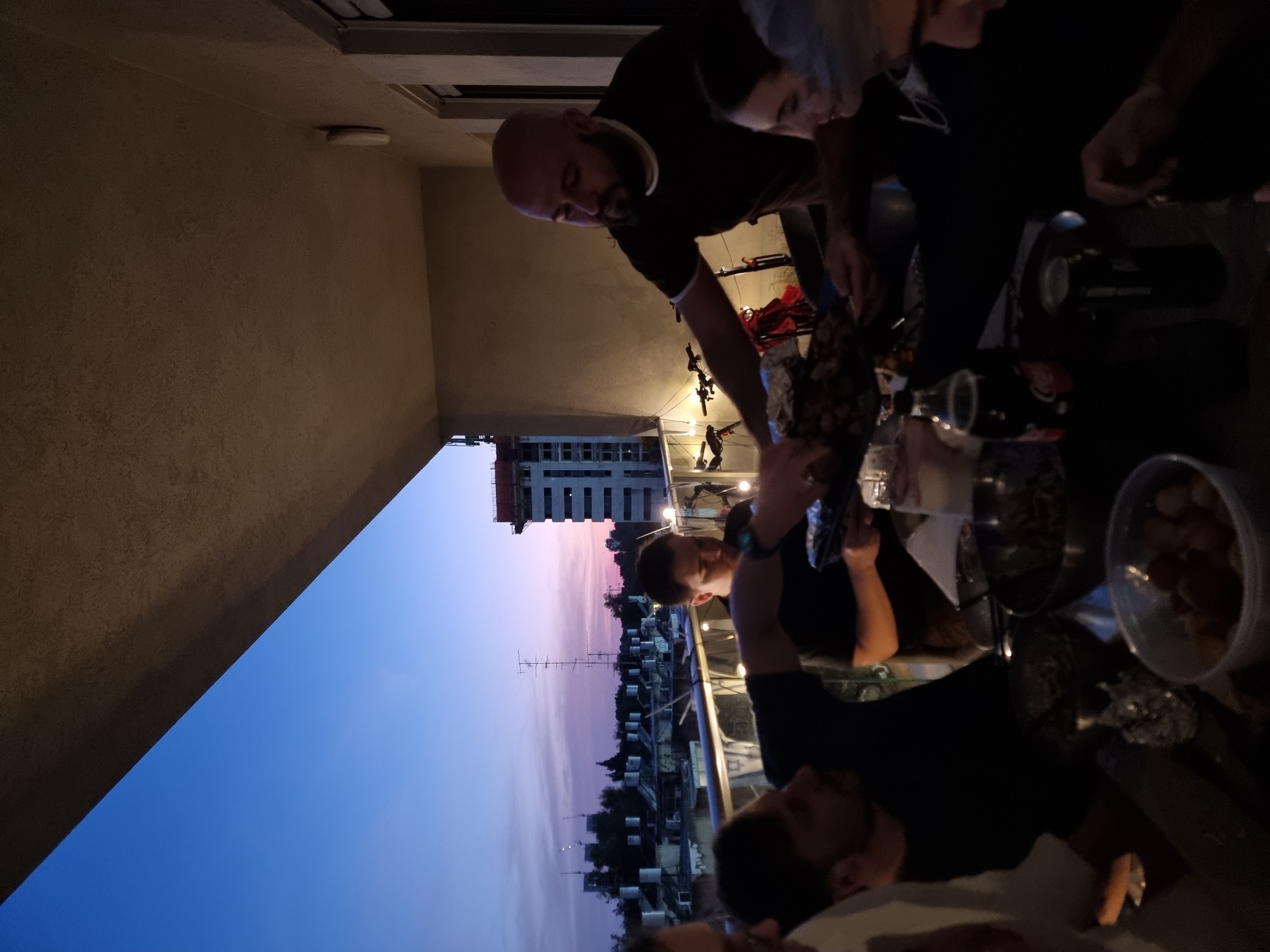 Several people are gathered around a table on a balcony in Ramat Gan, Israel, preparing food. The scene takes place during dusk, as the sky transitions from blue to pink. There is a building under construction visible in the background, along with an antenna and other rooftops. The table holds bowls, bottles, and cooking utensils. The people are wearing casual clothing.