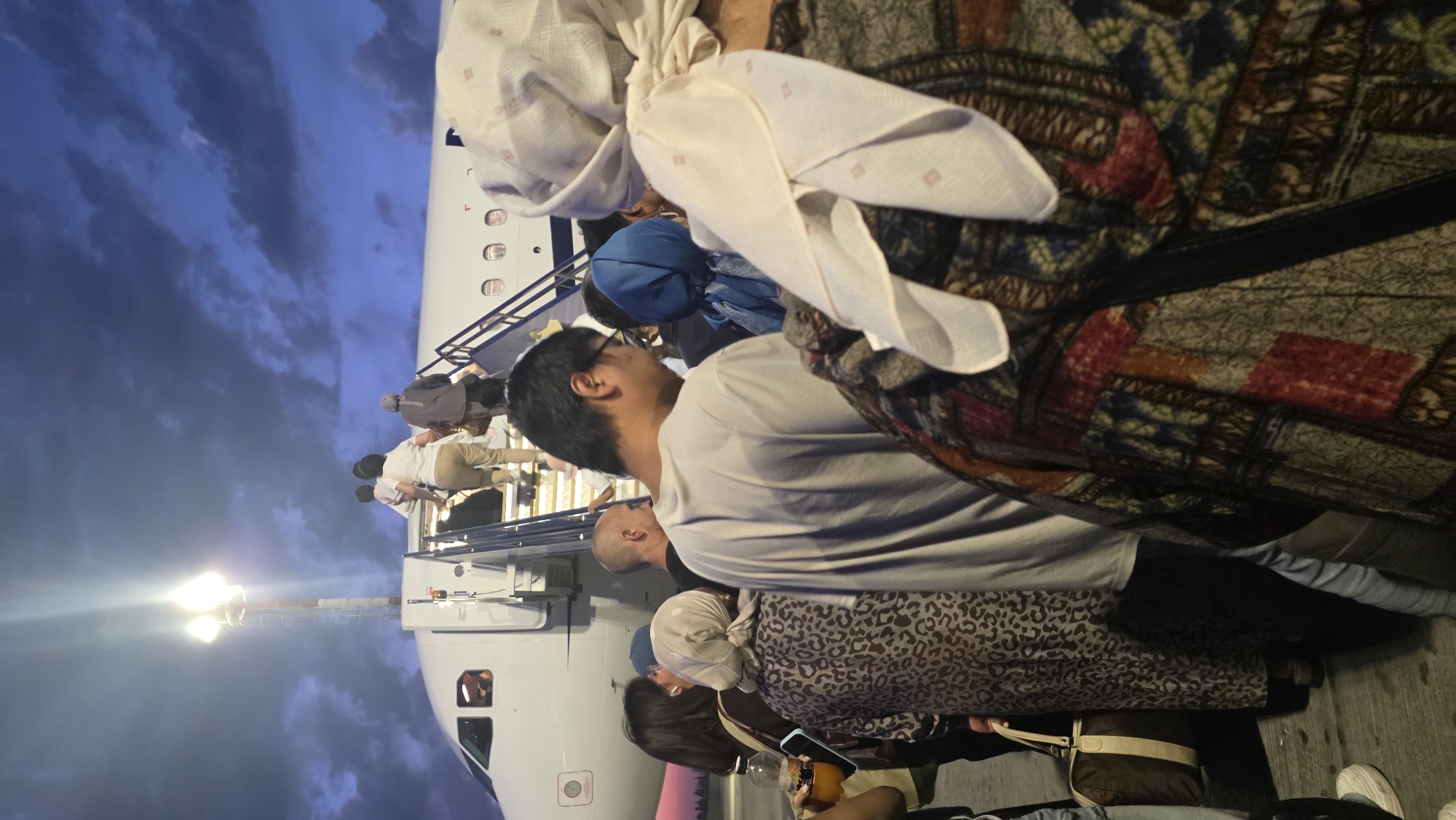 An evening view shows a group of people boarding an airplane via a staircase. The sky is dark blue with dramatic clouds, suggesting dusk or pre-dawn. A bright floodlight illuminates the top of the staircase. The people are dressed in casual clothing, with some women wearing headscarves. A suitcase is visible on the ground. The scene appears to be at an airport. The location in the city of түндүктө, Kyrgyzstan, is not discernible from the image.