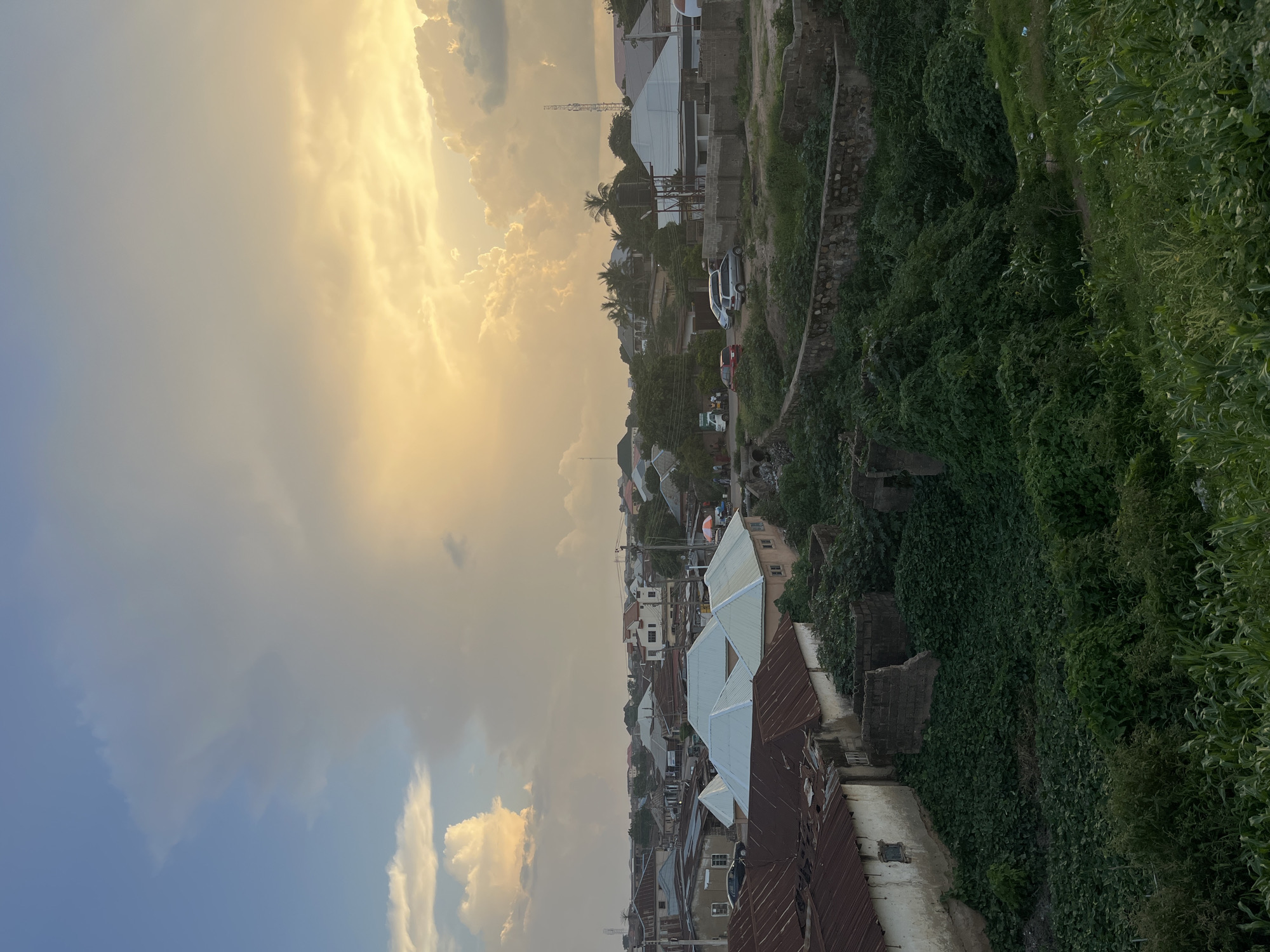 The image displays a panoramic view of a dense urban area in Jos, Nigeria, dominated by a dramatic sky. The sky is filled with a mix of bright, illuminated clouds, possibly during sunset or sunrise, casting a warm, golden glow on the scene. The lower part of the image reveals a collection of buildings with varied roofing materials, including corrugated metal and lighter colored tiles. Some structures appear newer with white facades, while others show signs of age with weathered roofs.
The settlement is built on a hilly terrain, evident from the way the buildings are positioned along a slope. The foreground is characterized by lush, overgrown greenery, with ivy and other plants covering walls and slopes, suggesting a humid climate and possibly a period of recent rainfall. A pathway or road winds through the settlement, with a few parked cars visible, indicating ongoing activity. The presence of a telecommunications tower in the distance provides a modern element to the otherwise rustic and densely packed residential landscape. The overall impression is one of a vibrant, living community nestled within a verdant, somewhat untamed natural environment, under a sky painted with the colors of dawn or dusk. No discernible text is present in the image.